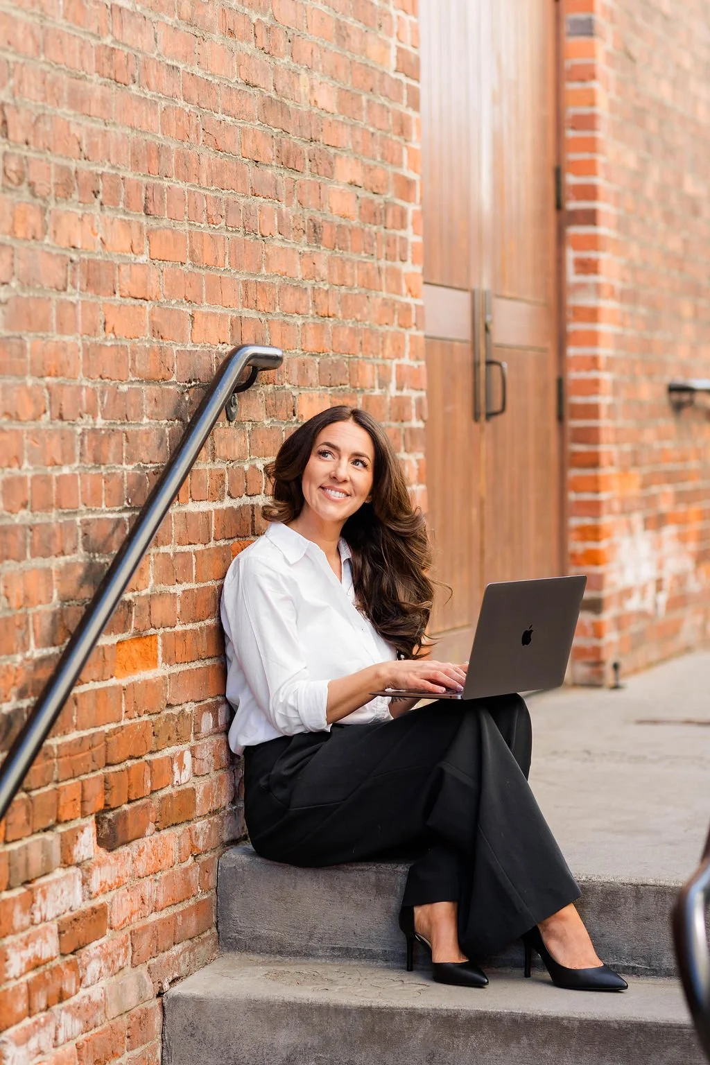 A woman sitting on a stair step outside a brick building, working on a laptop, smiling, dressed in a white button-up shirt and black pants, with black high heels.