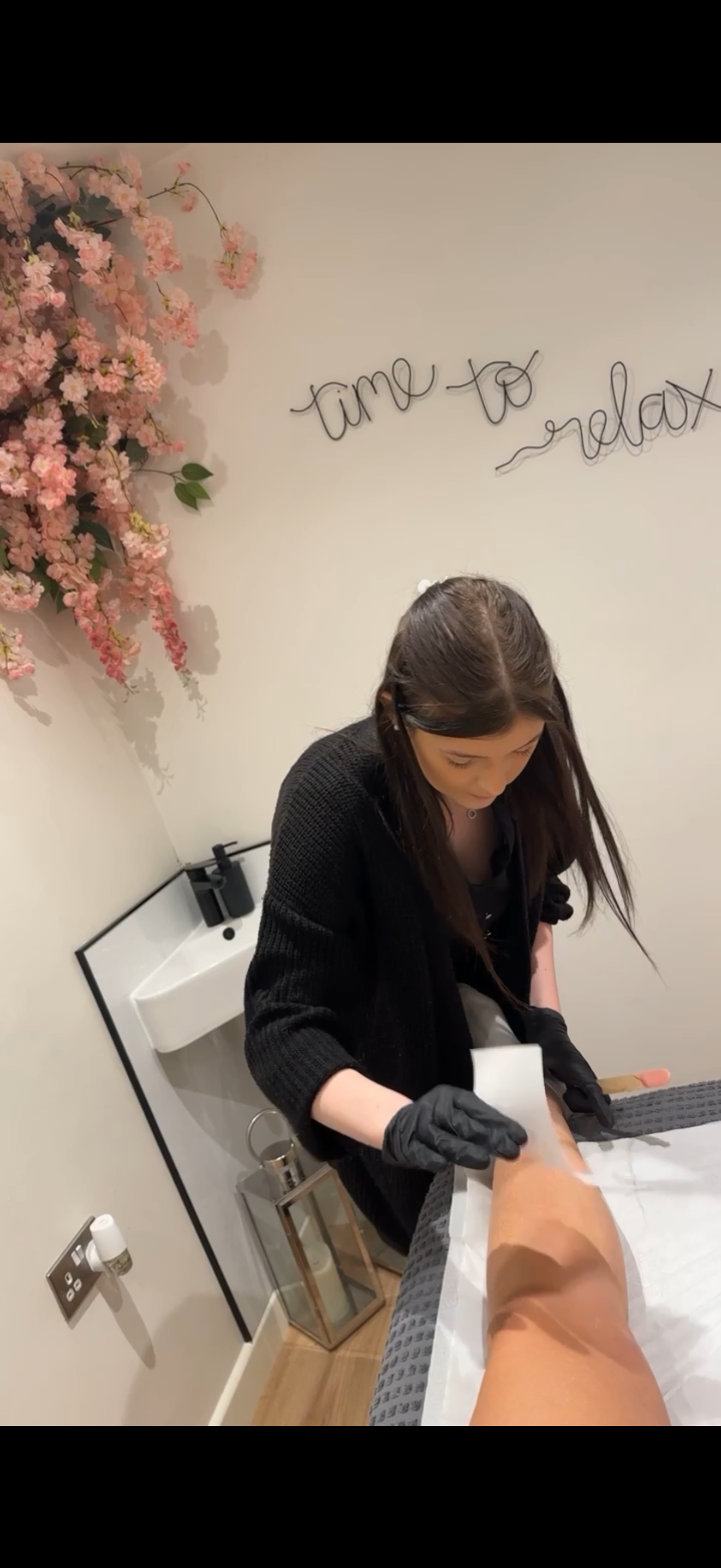 A woman giving a tattoo to a person on their arm, in a room decorated with pink flowers and a wall sign that reads 'time to relax'.