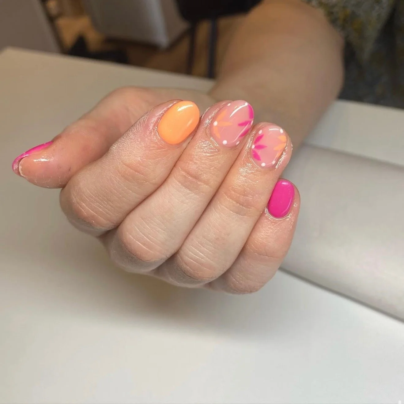 Close-up of a person's hand with colorful nail art. The thumb is painted in bright pink, the index finger has a peach-colored polish, the middle and ring fingers feature a clear base with pink, orange, and white flower and dot designs, and the pinky finger is painted vivid pink.