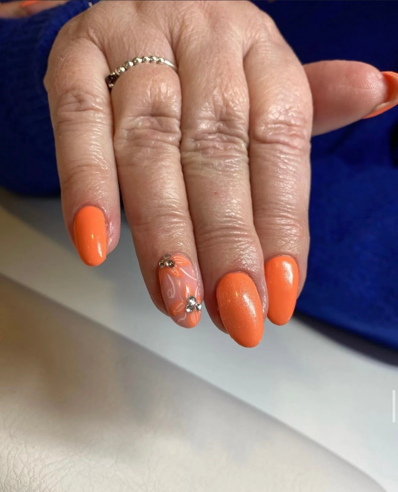 Hand with orange painted nails, one decorated with white swirl design and rhinestones, wearing a silver beaded ring.