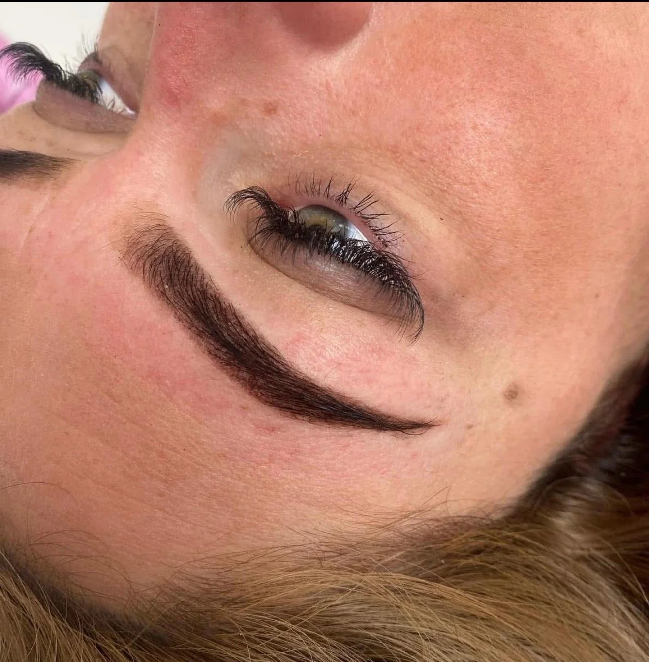 Close-up of a person's face with makeup, focusing on their eye, eyebrow, and part of their cheek. The person has dark, well-groomed eyebrows, long eyelashes, and light skin.