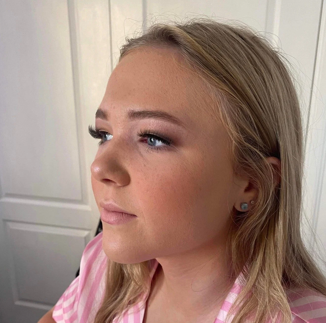 A close-up of a woman with blonde hair, blue eyes, and makeup, wearing a pink and white striped shirt, indoors with a white door in the background.