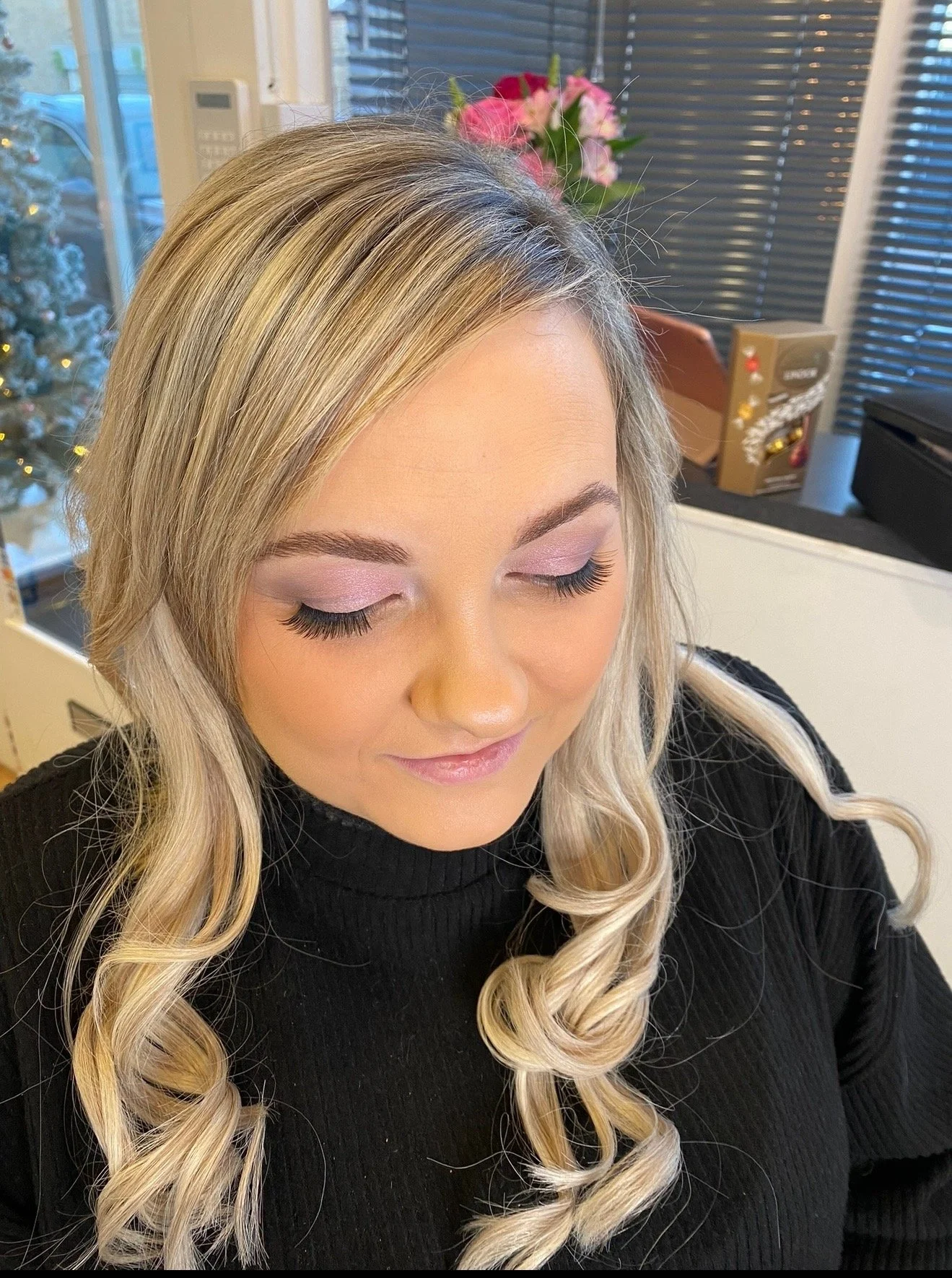 A woman with long blonde hair styled in loose curls, wearing light makeup with pink eyeshadow and pink lipstick, looking down with her eyes closed in a room decorated for Christmas with a small Christmas tree, pink flowers, and holiday decorations in the background.