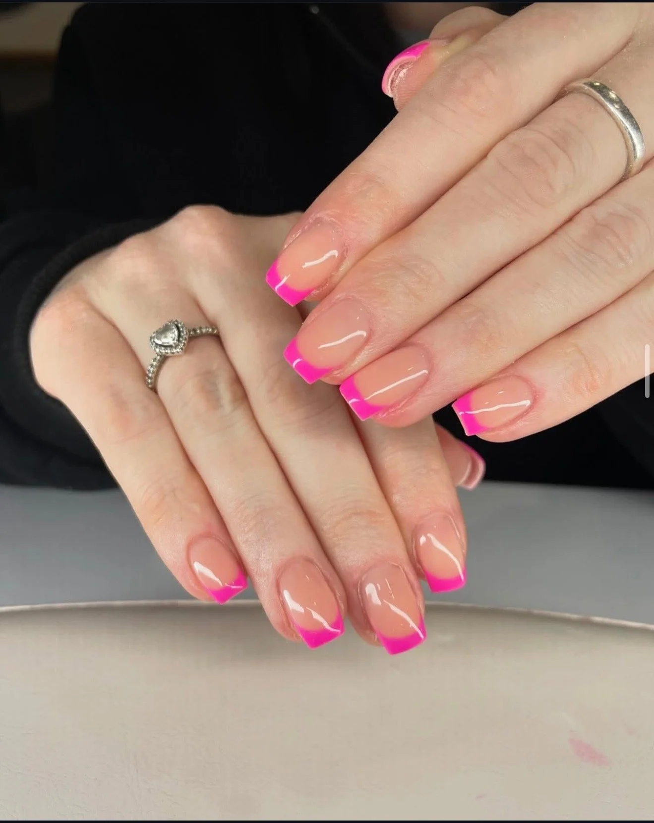 Close-up of a person's hands with pink French manicured nails, wearing a silver ring with a heart-shaped stone on one finger and a simple silver band on another finger.