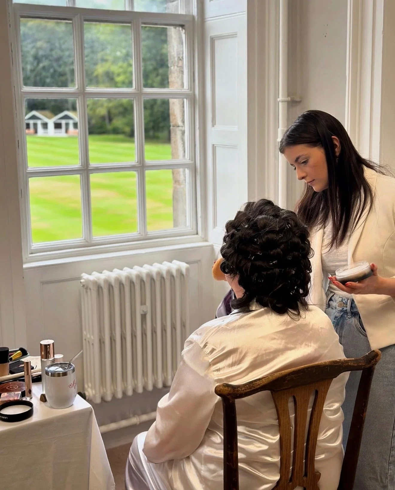 A woman with dark hair applying makeup to another woman with curly hair inside a room with white walls and a large window showing a green outdoor area.