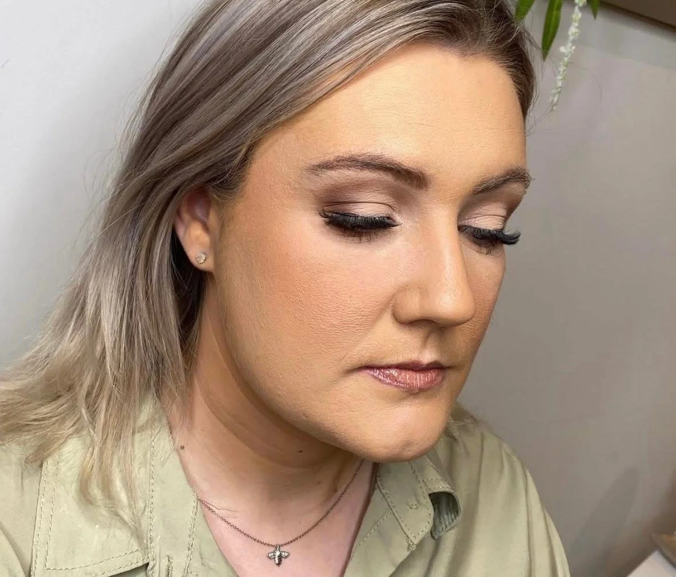 Close-up of a woman with blonde hair, eyes closed, wearing makeup, a small earring, a necklace with a cross pendant, and a light-colored shirt, in front of a neutral background.