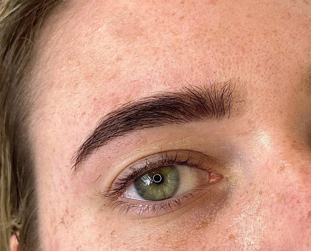 Close-up of a person's eye with well-groomed eyebrow and light skin, showing green iris and minimal makeup.
