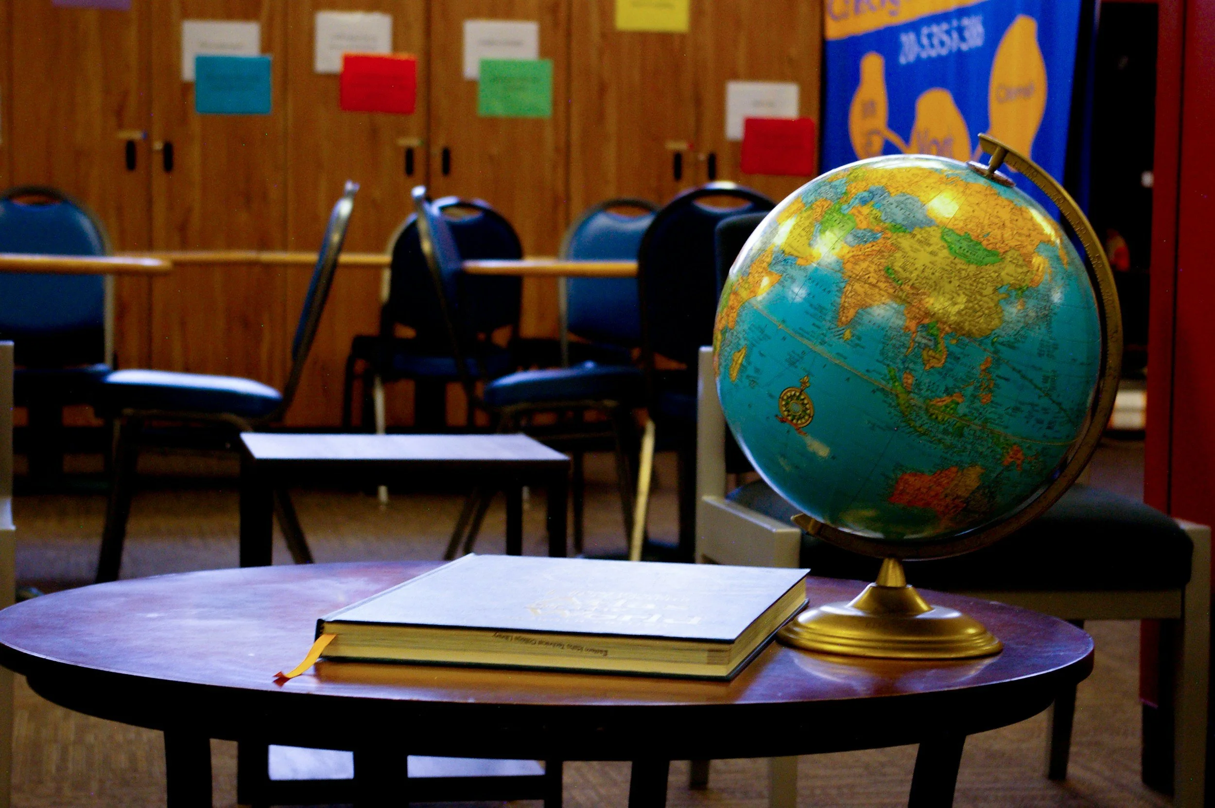 A classroom setting with a globe and a closed book on a round wooden table. In the background, there are chairs and colorful posters on a wooden wall.
