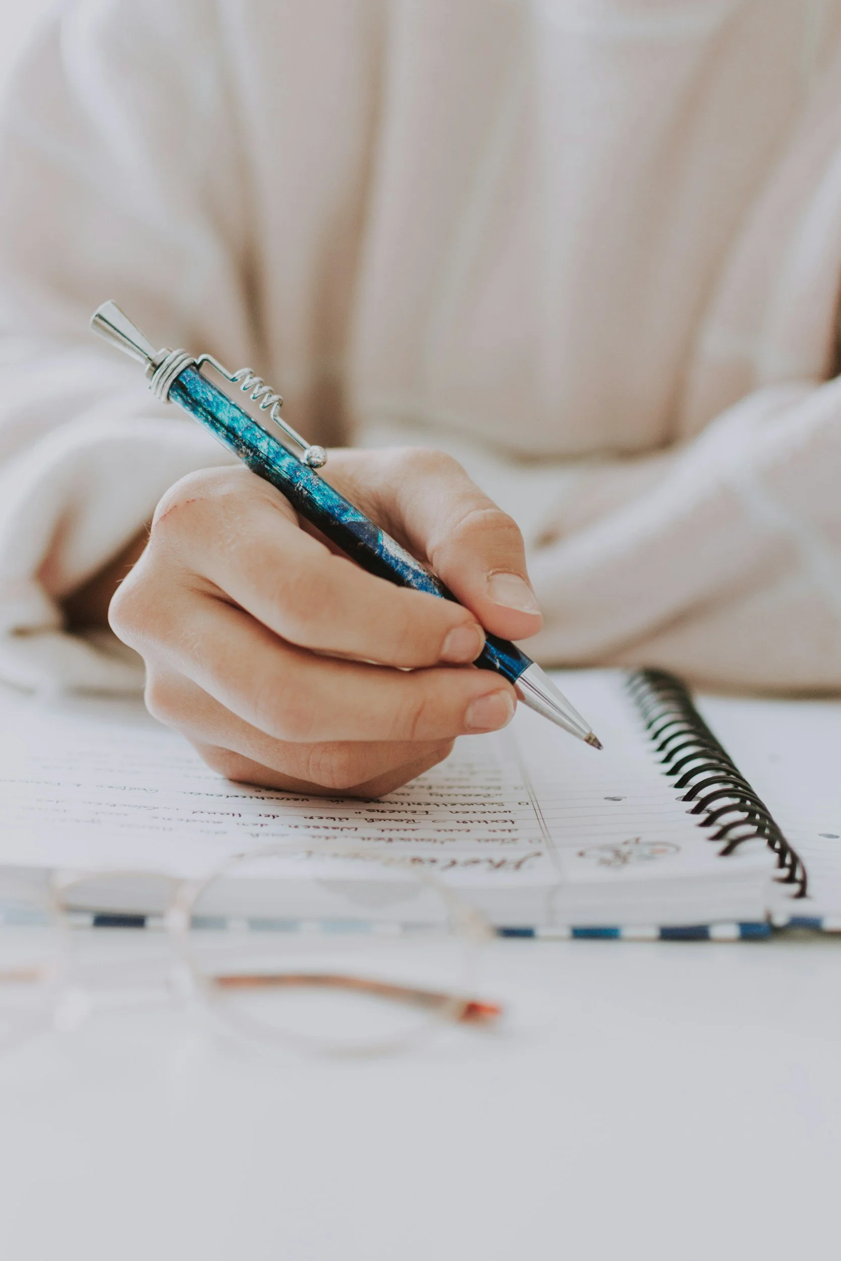 A person writing in a spiral notebook with a blue and silver pen, on a white surface with glasses nearby.