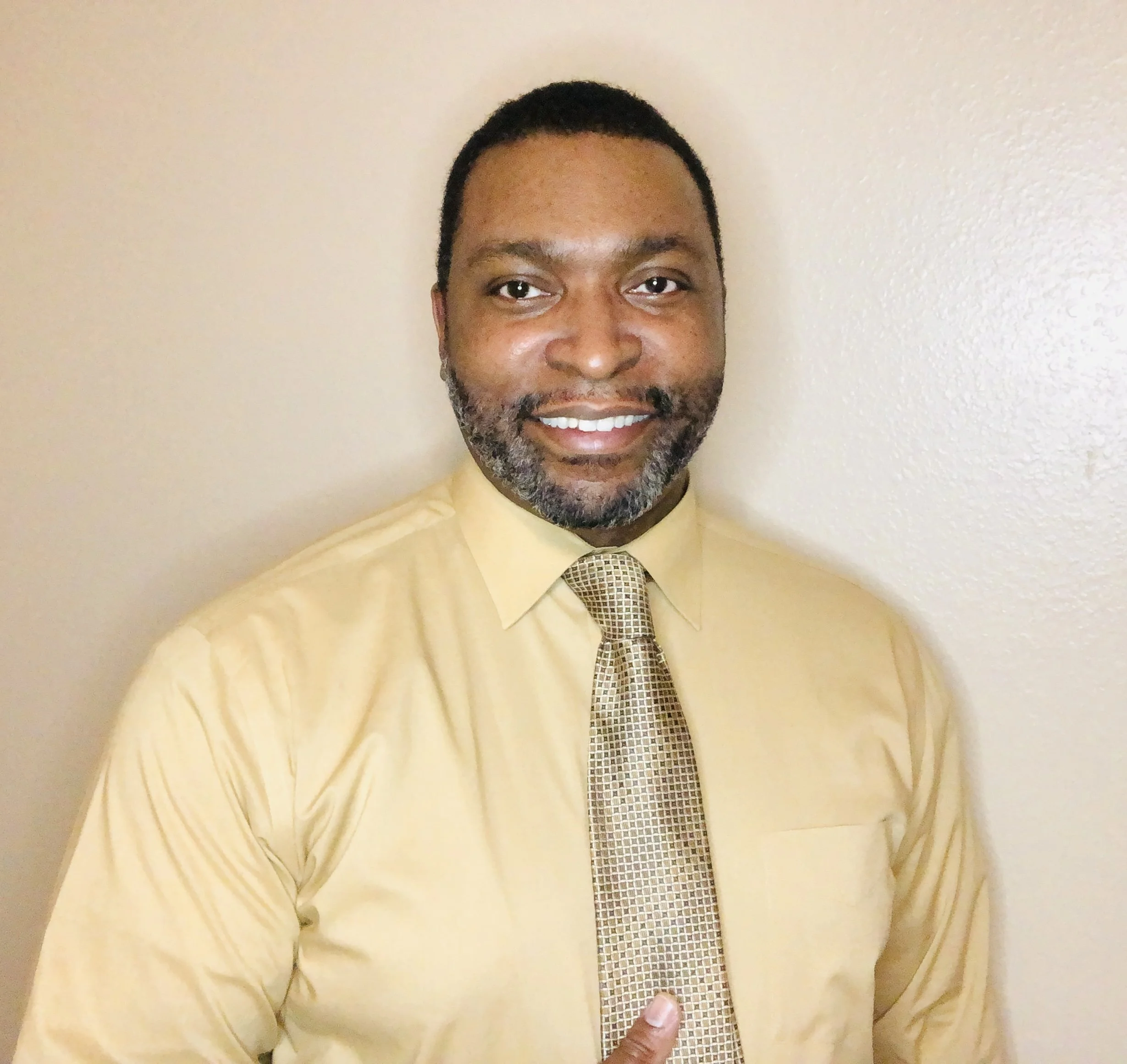 A smiling African American man with short black hair, wearing a yellow dress shirt and a patterned gold tie, standing against a beige wall.