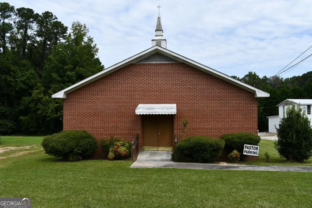 Red brick church building with a white steeple, surrounded by green bushes and grass, and a sign that reads "Pastor Parking".