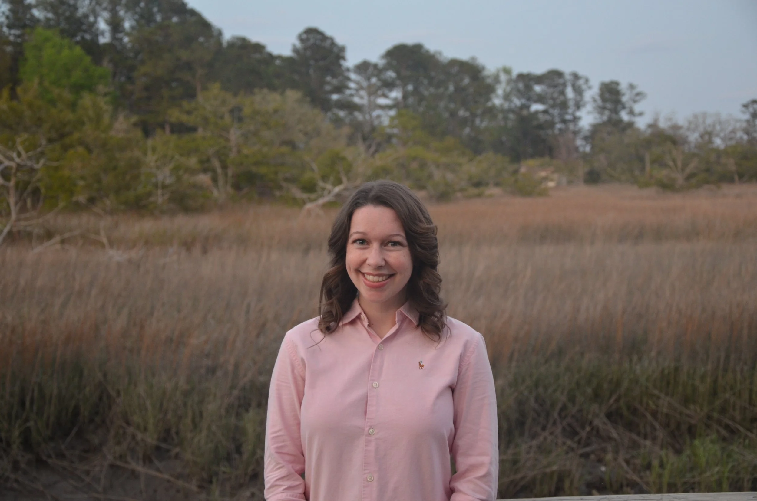 A woman in a pink button-up shirt standing outdoors in a grassy field with trees in the background.
