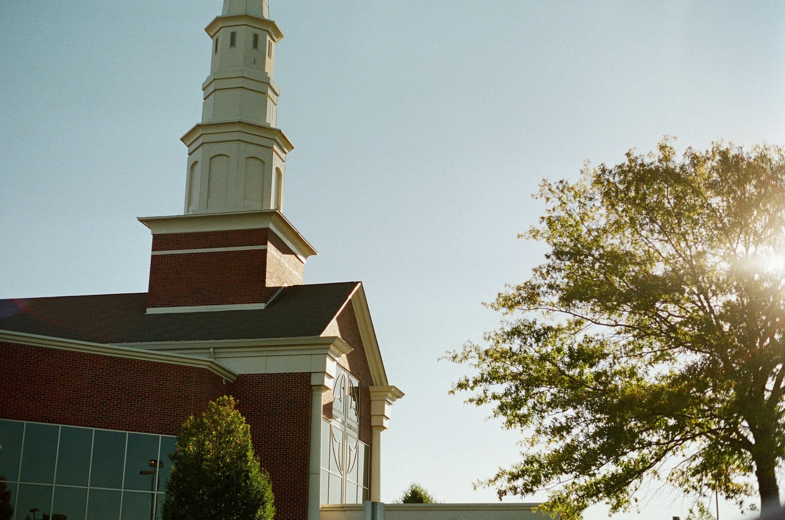 A brick building with a tall white steeple and a clock, partially obscured by trees, under a clear sky with sunlight.