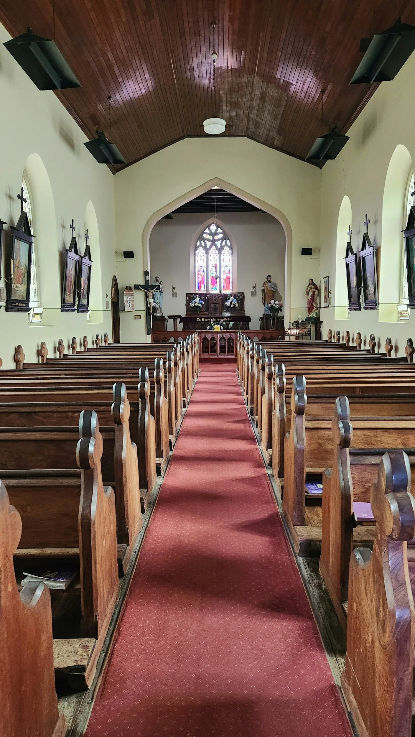 Inside a church with wooden pews and a red carpet aisle leading to the altar. The altar has religious statues and stained glass windows. The walls have framed religious artwork and the ceiling is made of wood panels with hanging speakers.
