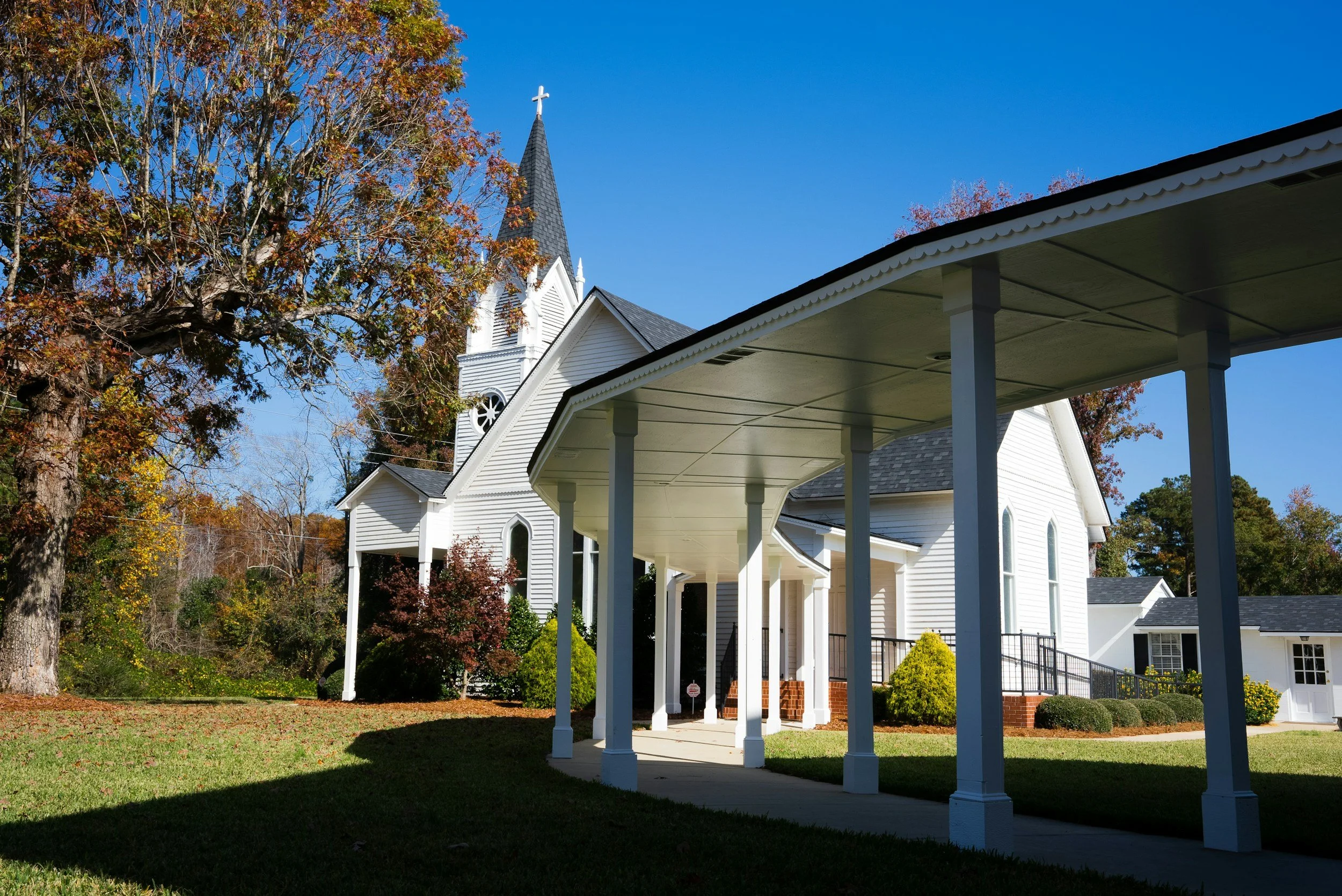 View of a white church with a tall steeple topped by a cross, surrounded by autumn trees with colorful leaves, against a clear blue sky, and a covered walkway supported by white columns.