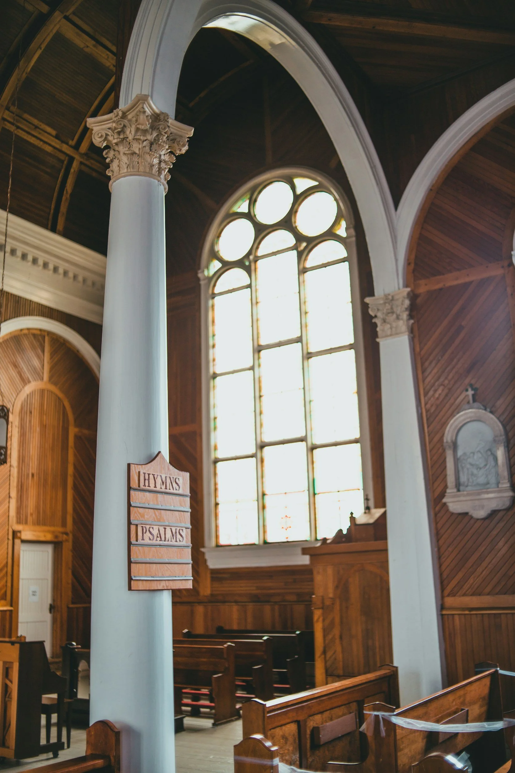 Interior of a church with tall arched stained glass windows, wooden pews, and decorative columns with ornate capitals.