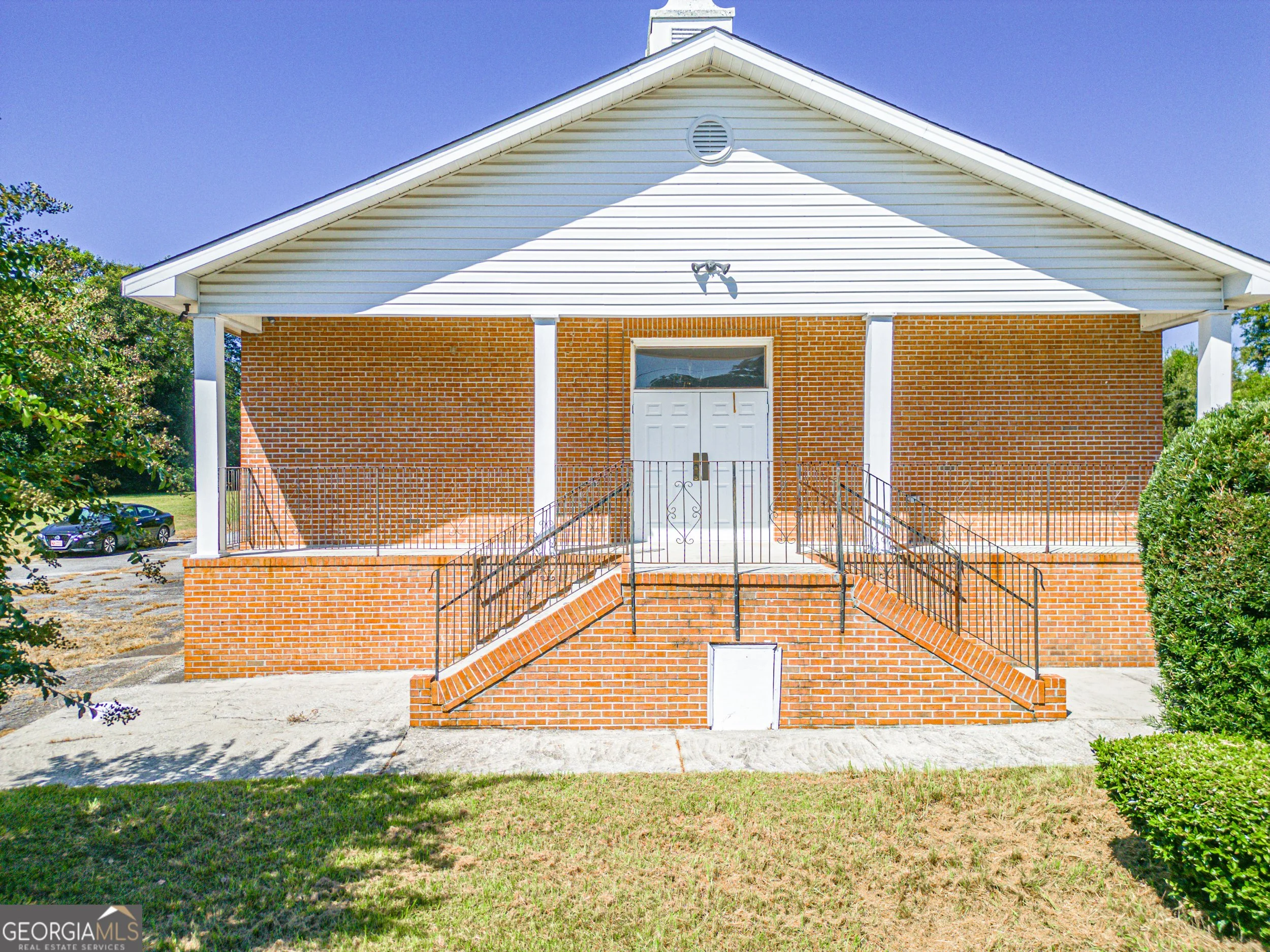 Front view of a residential building with brick lower walls, white siding upper walls, a front door accessed by stairs, and a small bell tower at the top of the roof. The building has a fenced front yard, trees surrounding it, and a driveway with parked cars on the side.