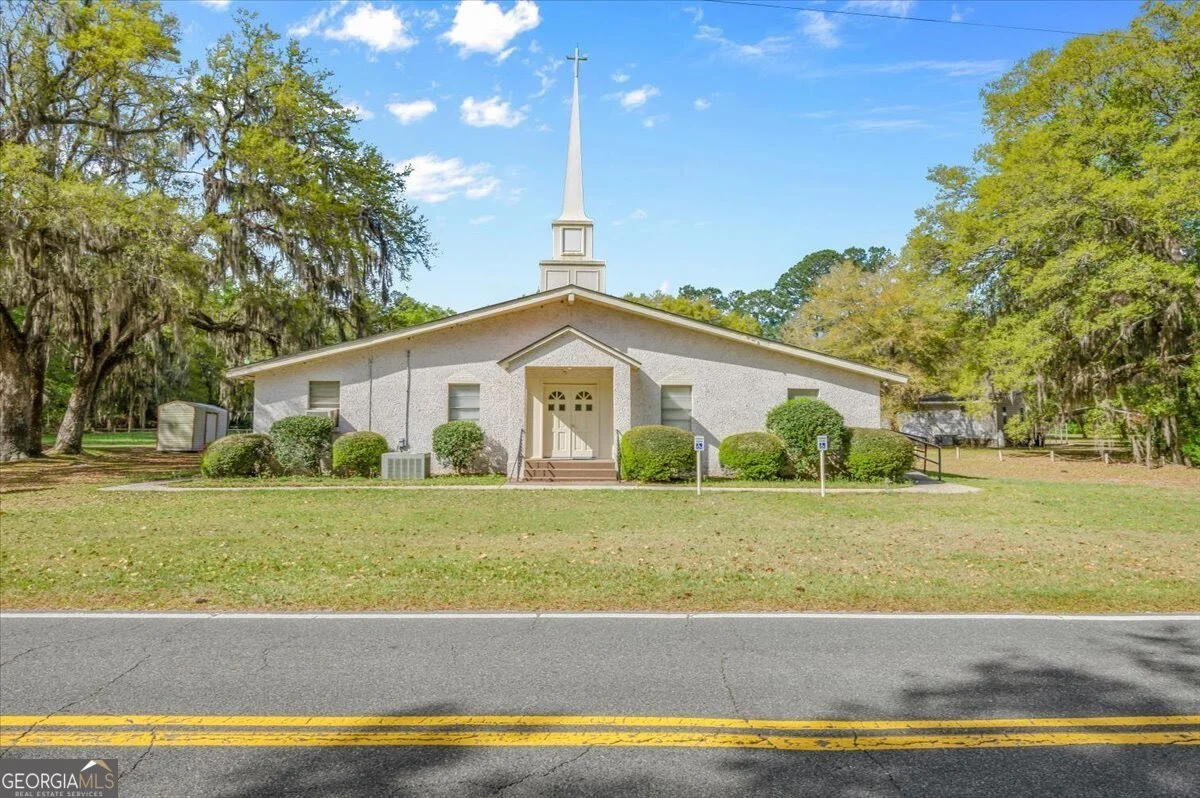 A church building with a tall steeple and a white exterior, surrounded by trees and bushes, with a parking lot and street in the foreground.