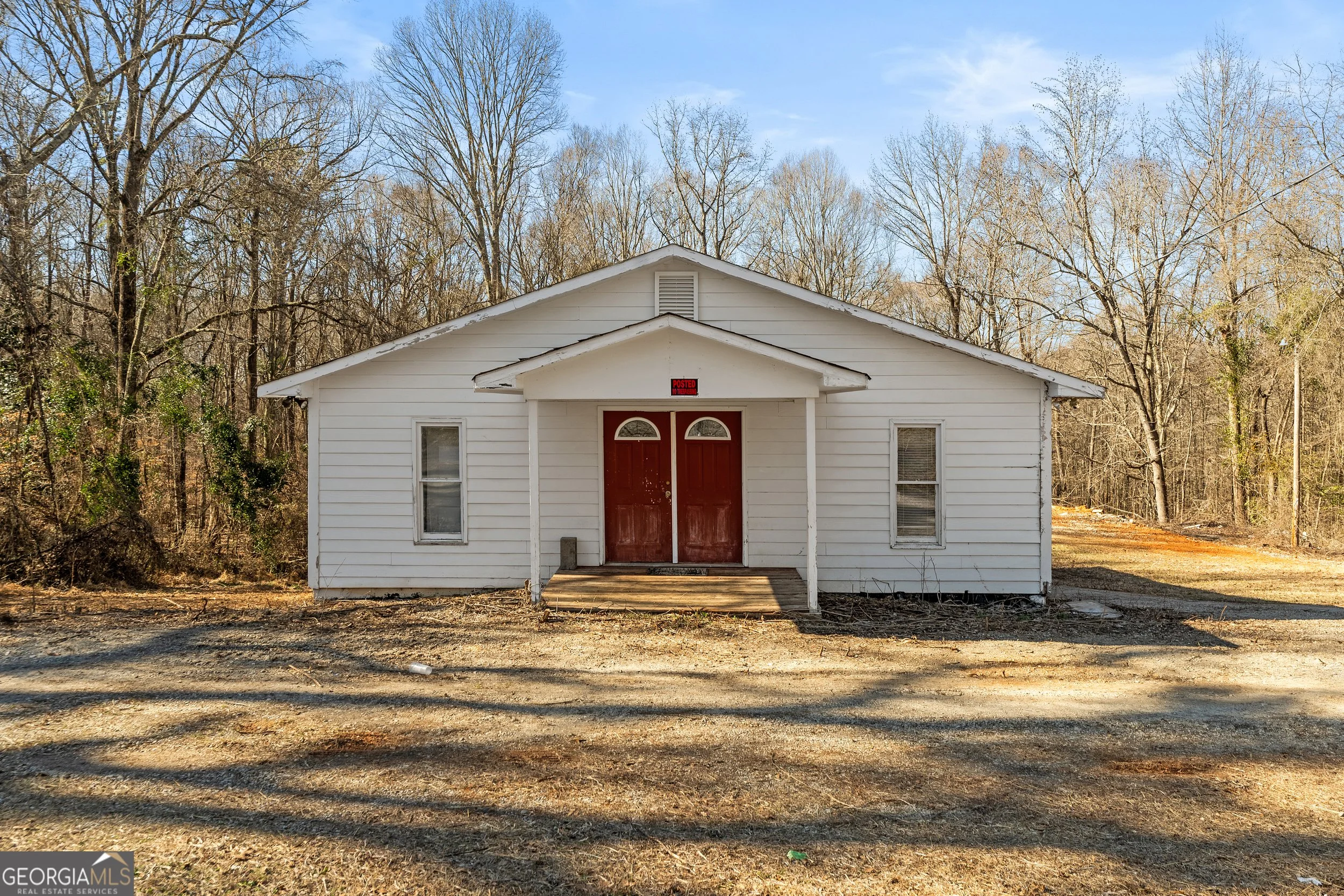 A small, white house with a red double door, situated in a wooded area with leafless trees and a dirt ground under a blue sky.