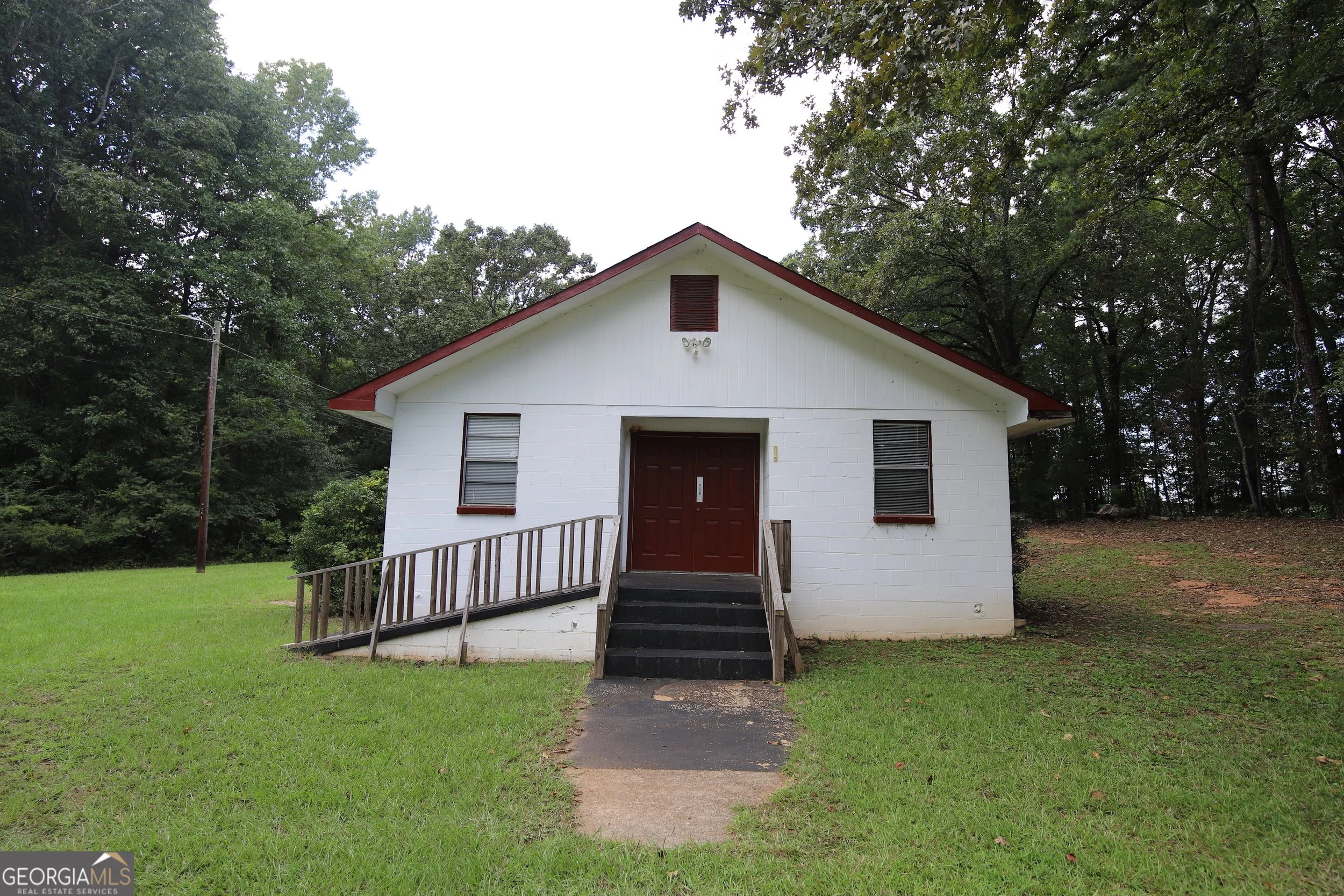 A small white house with a red roof, front porch, and roller shades in the windows, situated on green grass and surrounded by trees.
