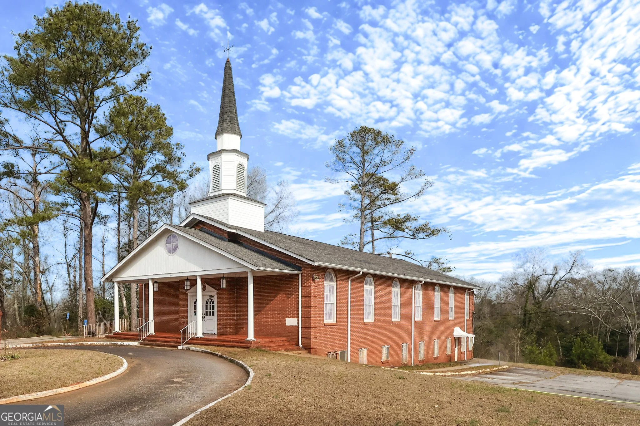 A white church with a brick foundation and a tall steeple, surrounded by trees and a driveway, under a sky with scattered clouds.