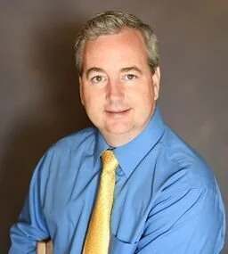 Portrait of a man with light skin, gray hair, wearing a blue shirt and yellow tie, against a neutral background.
