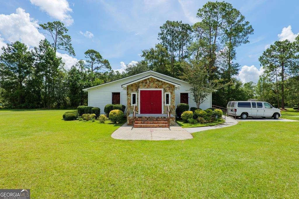 White building with a red door, surrounded by bushes and trees, with a parked white van nearby, under a partly cloudy sky.
