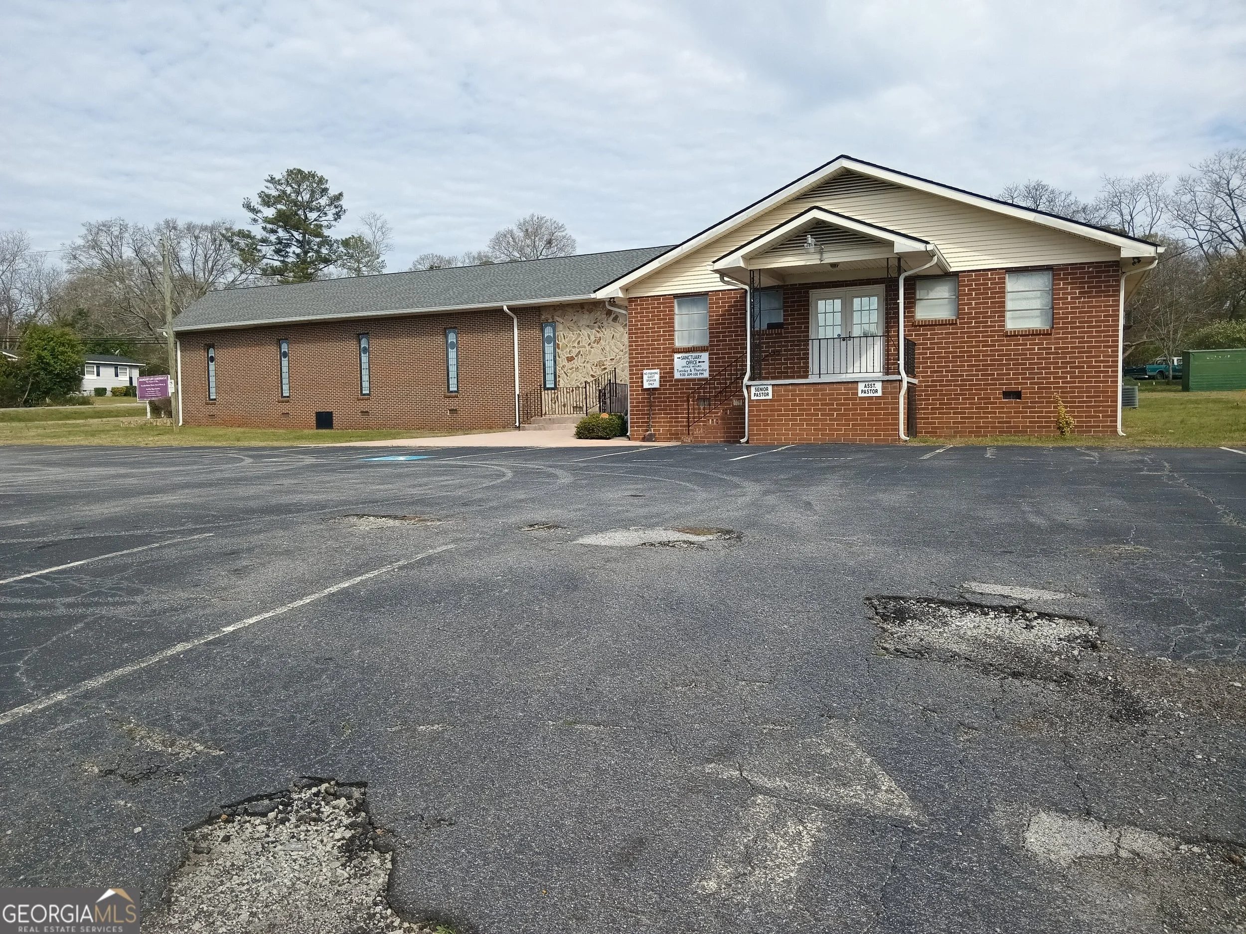 Parking lot in front of a church building with signs indicating office hours and pastoral staff, with trees and cloudy sky in the background.