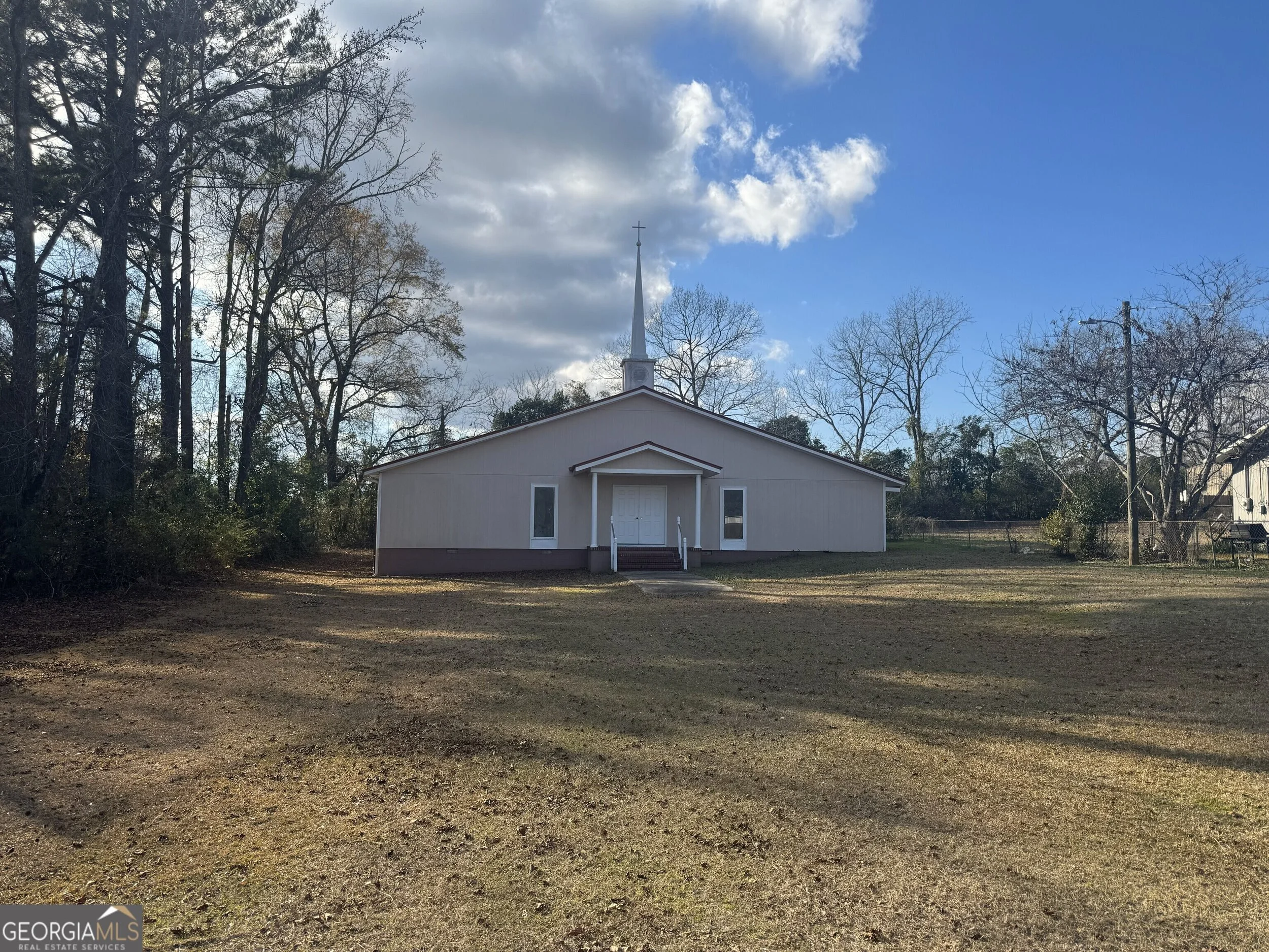 A small church with a white steeple and cross on top, surrounded by leafless trees and an open dirt area in front, under a partly cloudy blue sky.
