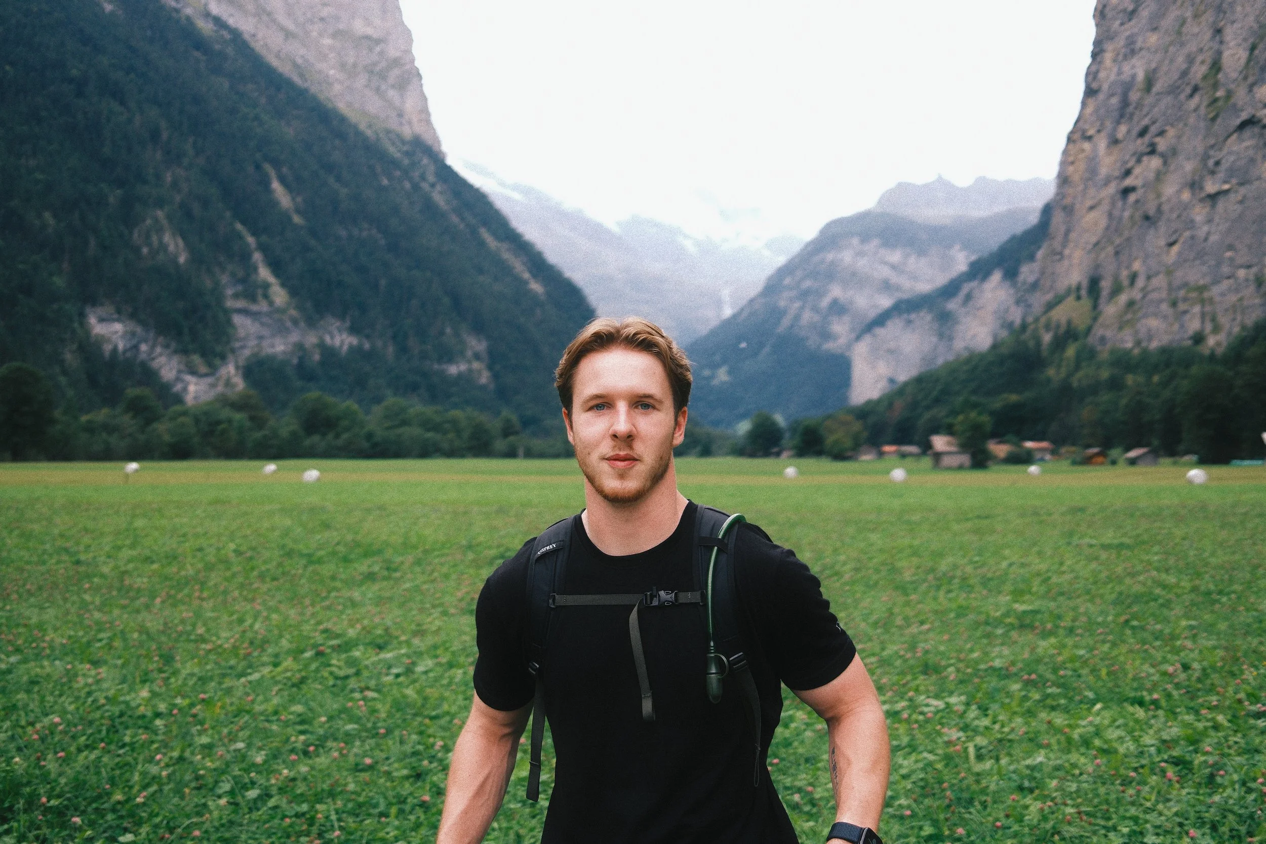 A young man with a backpack standing in a green field with mountains in the background.