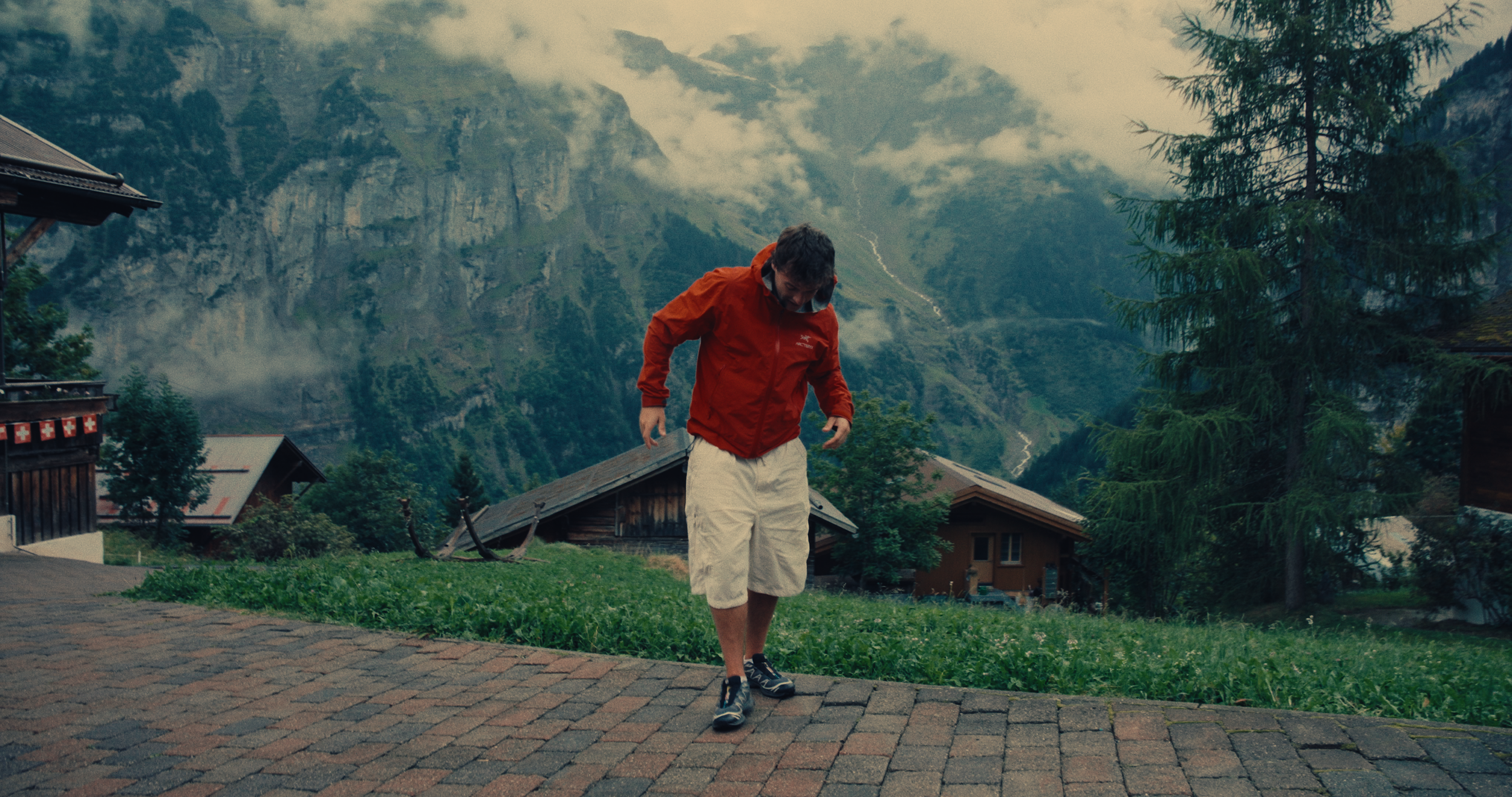 A man in a red jacket and white shorts walking on a brick pathway in a mountainous village.