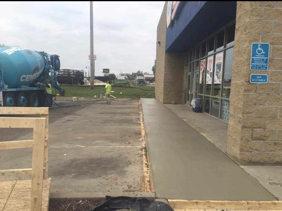 Construction workers pouring concrete for a sidewalk outside a commercial building with storefront windows. A cement truck is on site, and there are parking spaces and signage for handicapped parking.