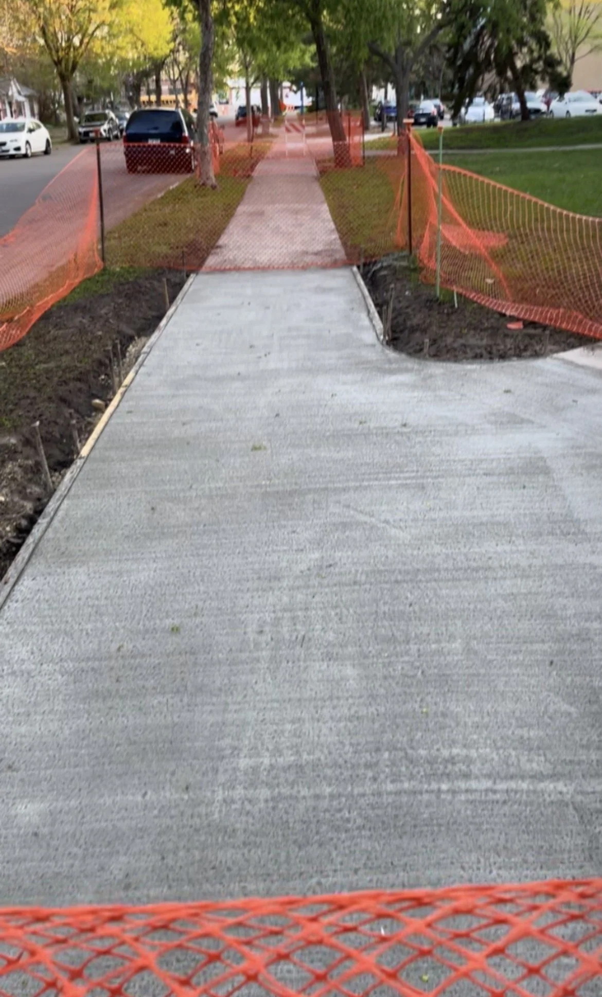 Sidewalk under construction with orange safety fencing and a new concrete pathway, trees and parked cars in the background.