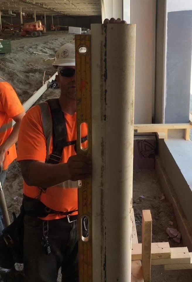 A construction worker wearing a white hard hat, sunglasses, and an orange shirt holds a level tool on a construction site.