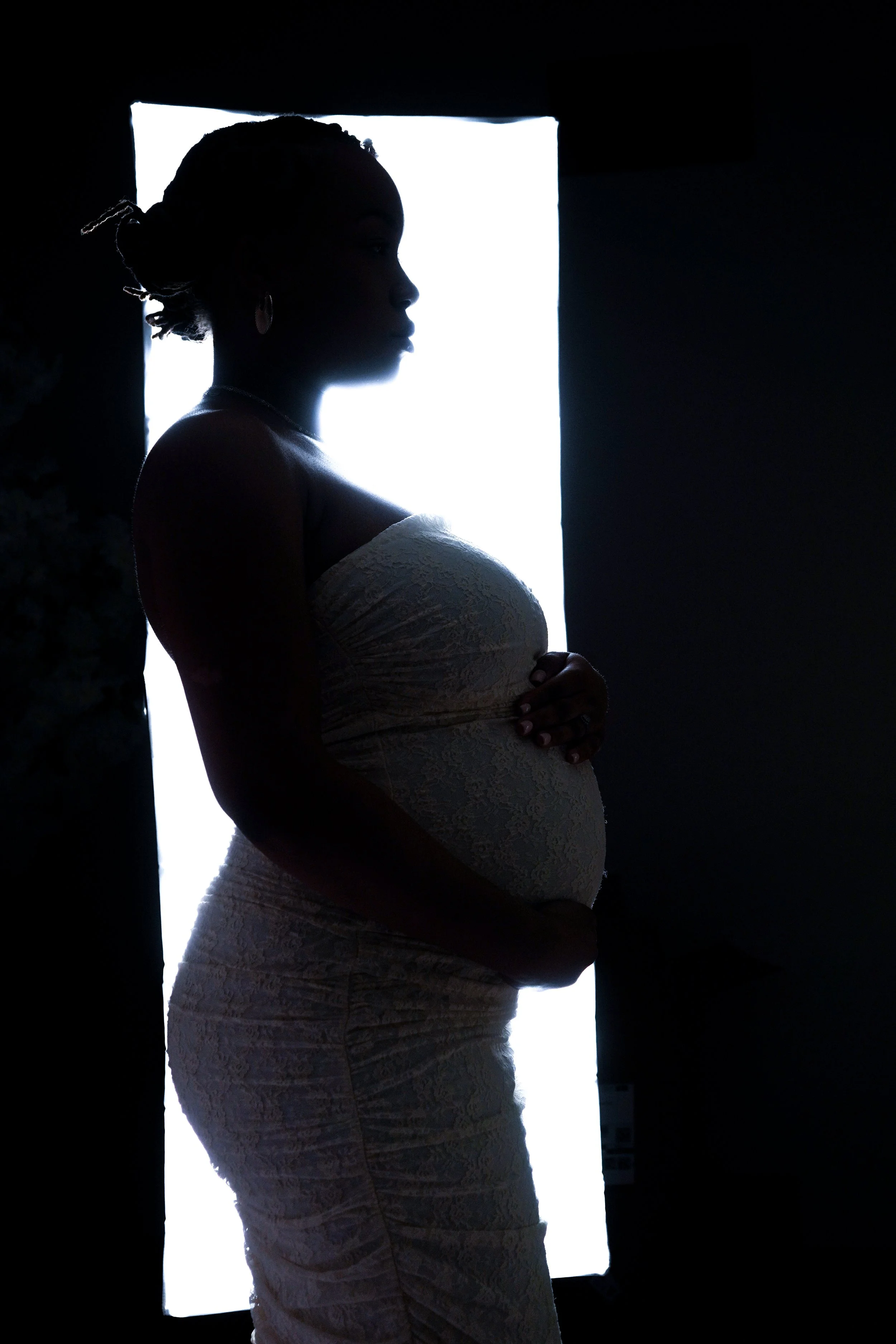 Silhouette of a pregnant woman in a lace dress standing in front of a bright window, holding her belly.