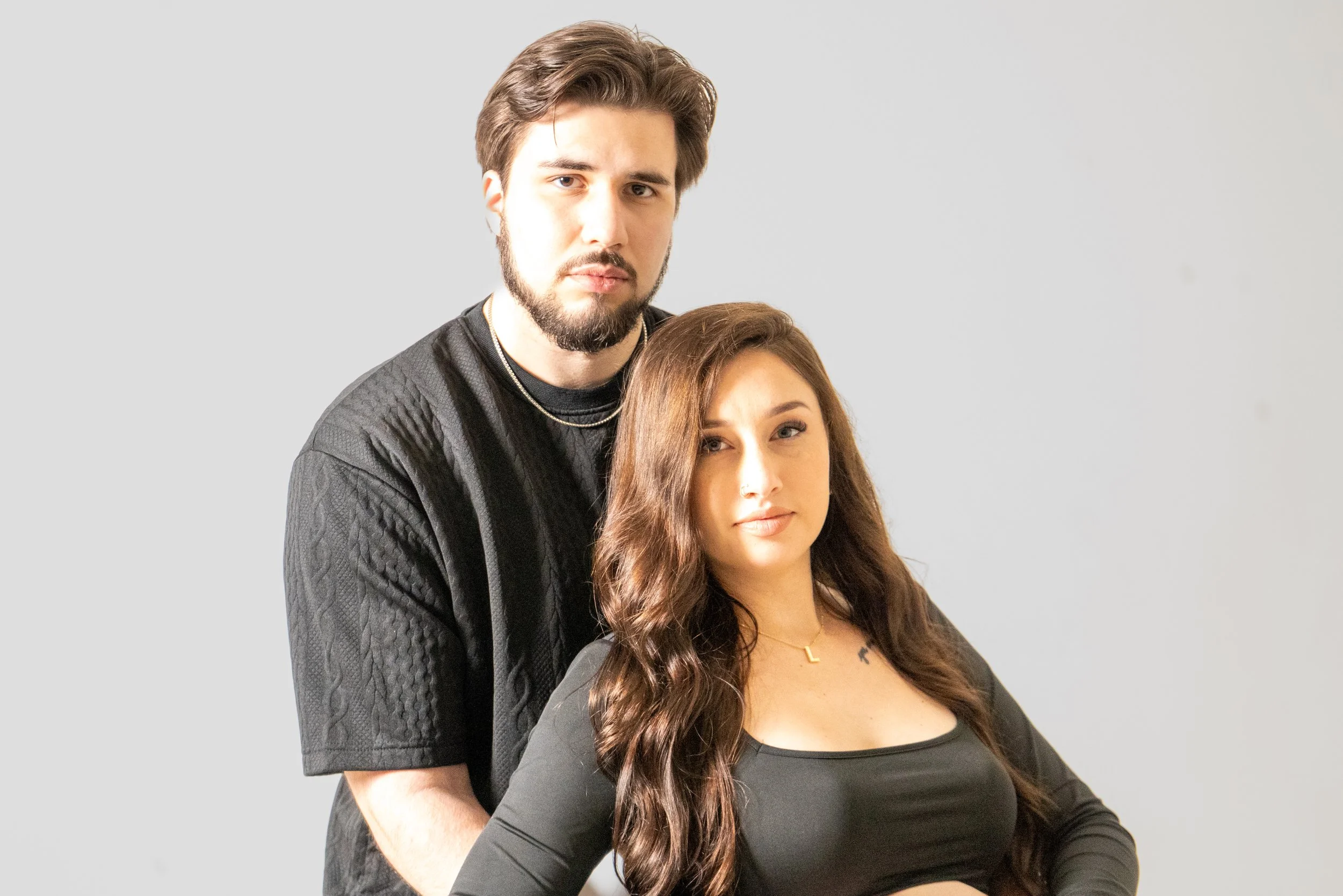 A man with dark hair and beard standing behind a woman with long wavy brown hair, both looking at the camera against a plain light gray background.