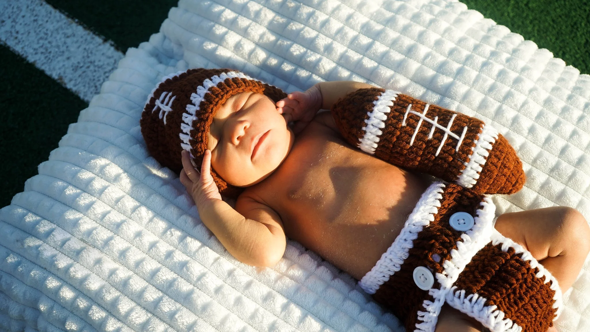 A newborn baby dressed as a football player, wearing a brown and white knitted football hat and matching shorts, is lying on a white quilted blanket outdoors, with one hand touching its face and the other relaxed beside.