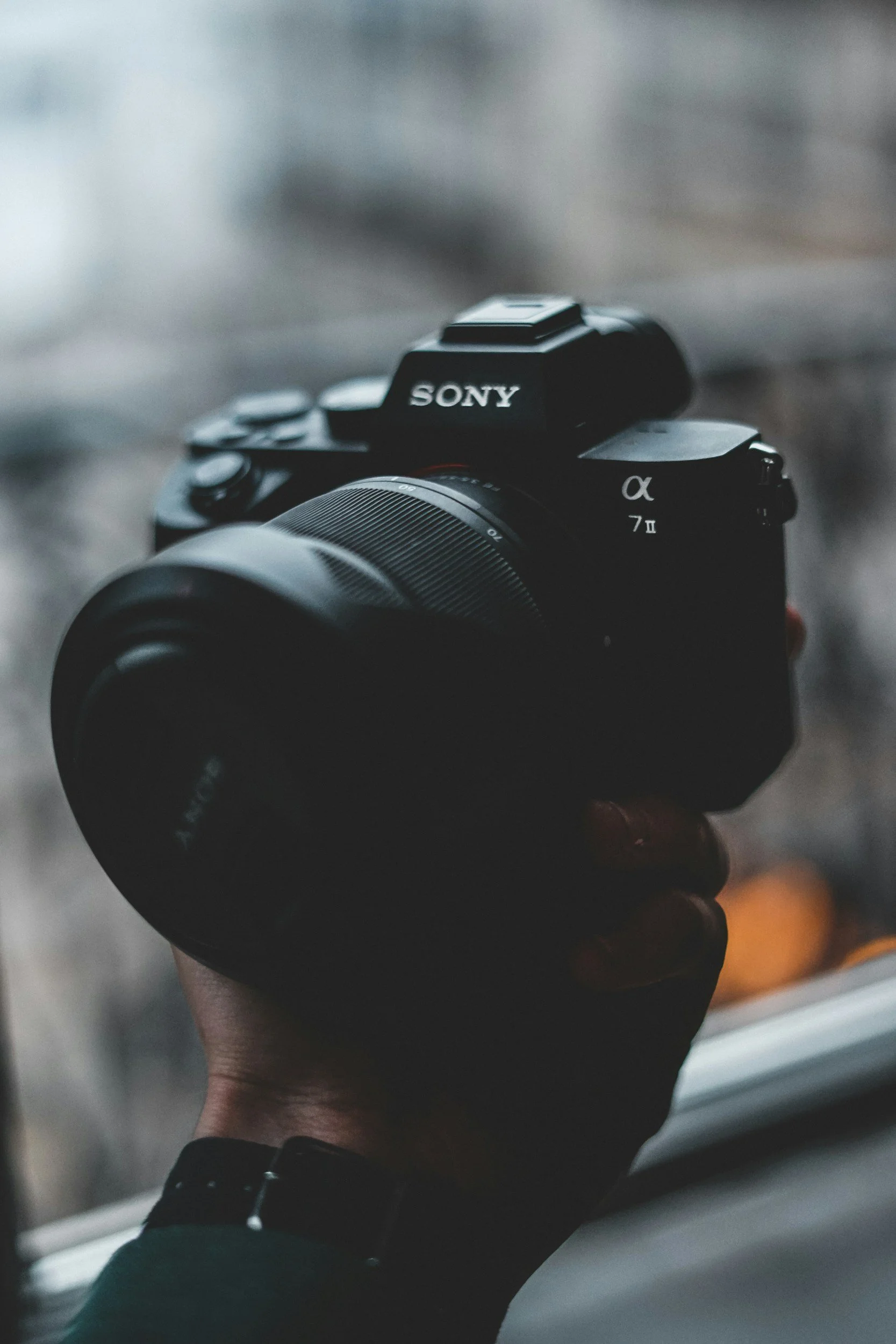 A person holding a black Sony camera with a zoom lens, the person's wrist is visible with a watch, and they are near a window with a blurry outdoor background.