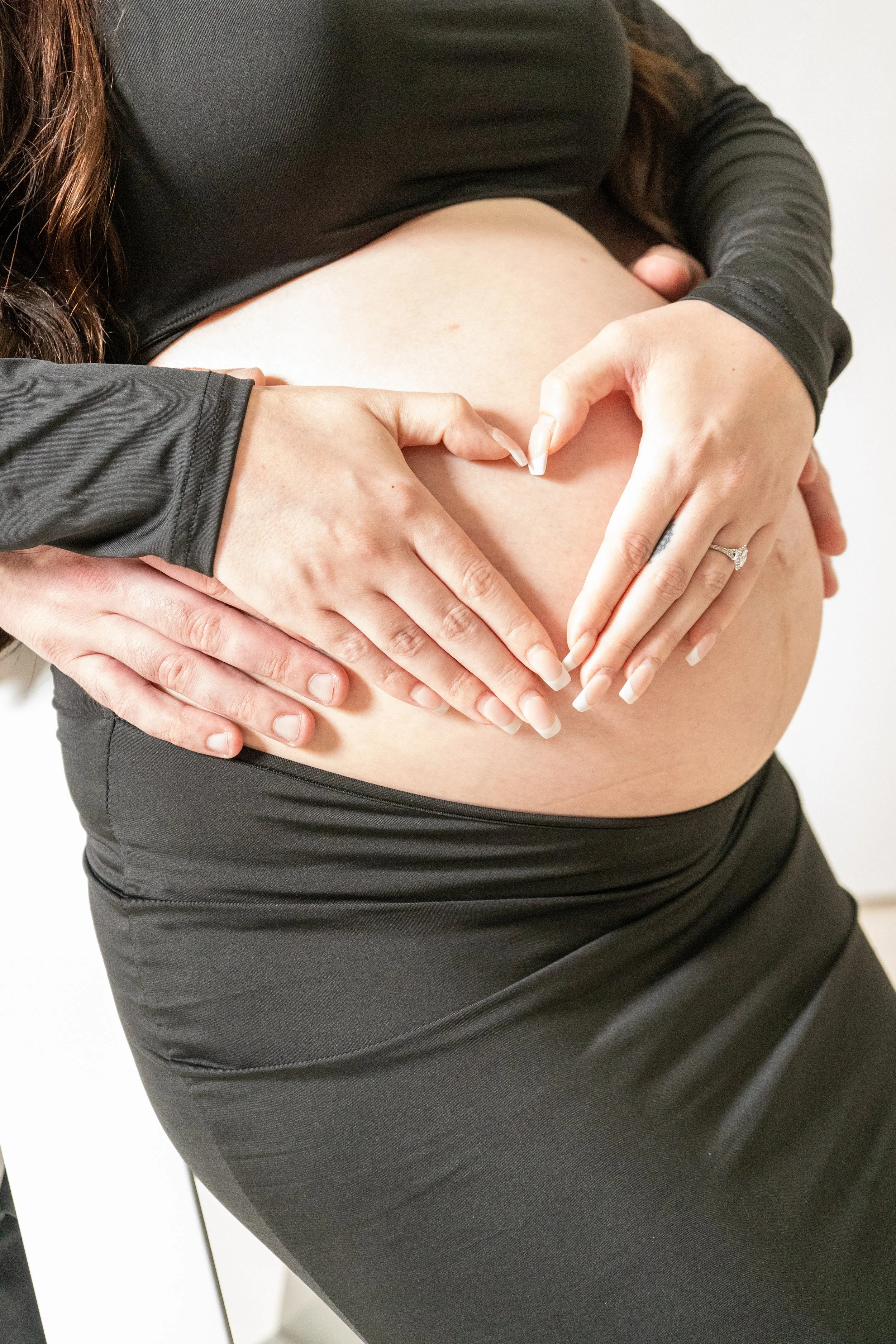 A pregnant woman making a heart shape with her hands on her belly, wearing a black top and black skirt.