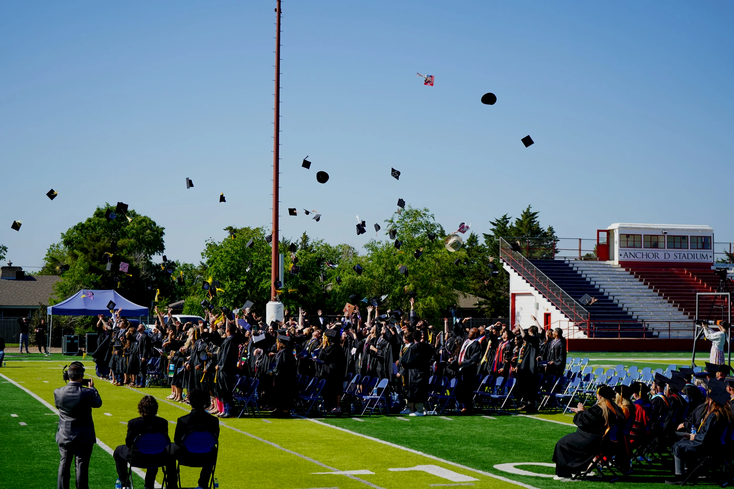 Graduates in caps and gowns throwing caps in the air during a graduation ceremony at a stadium field on a sunny day.