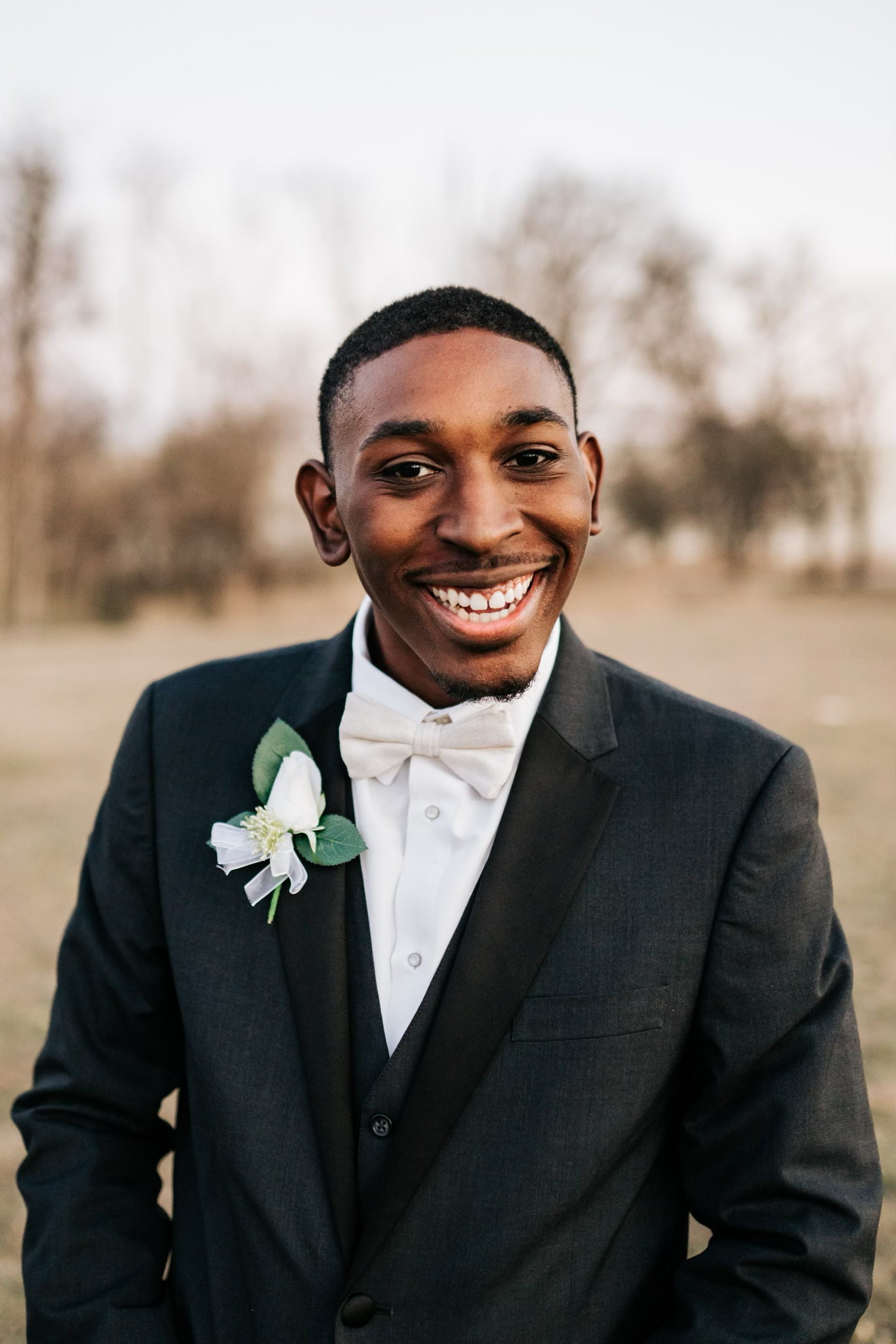 A young man in a formal black tuxedo with a white bow tie and boutonniere, smiling outdoors with trees in the background.