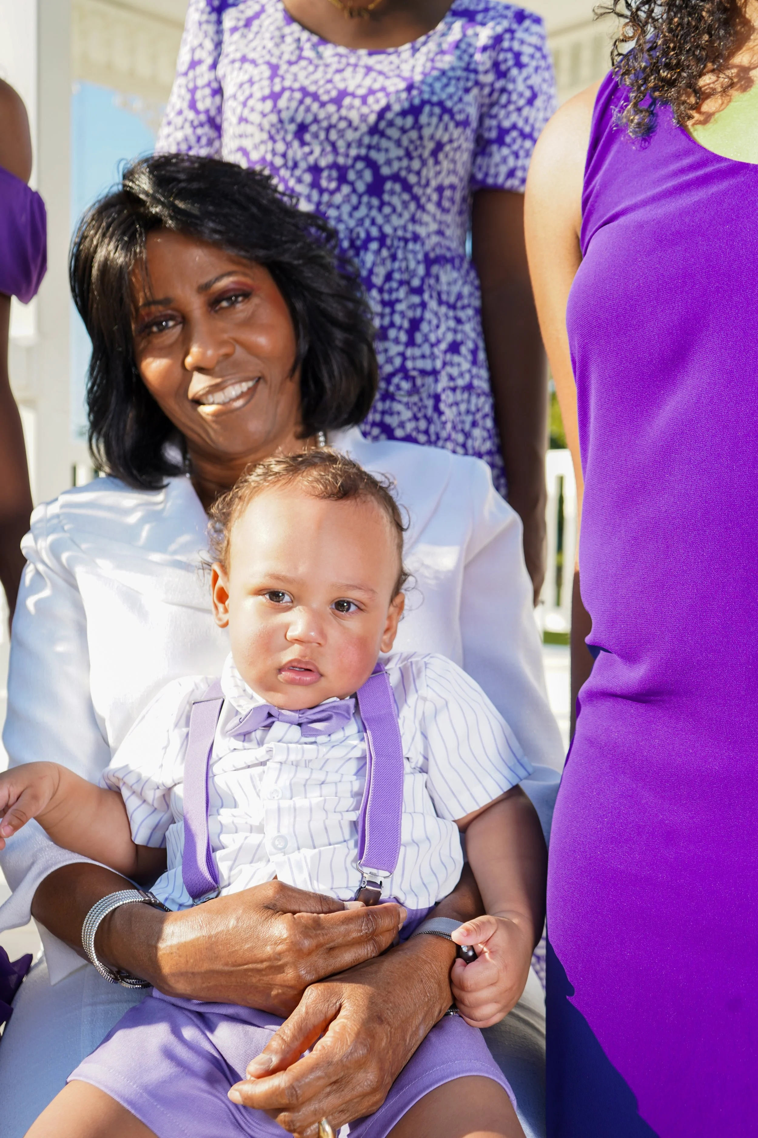 A woman with black hair, wearing a white jacket, sitting outdoors, holding a young boy on her lap. The boy has light skin, curly hair, is dressed in a white striped shirt with purple suspenders and a bow tie, and looks slightly confused. Partially vi
