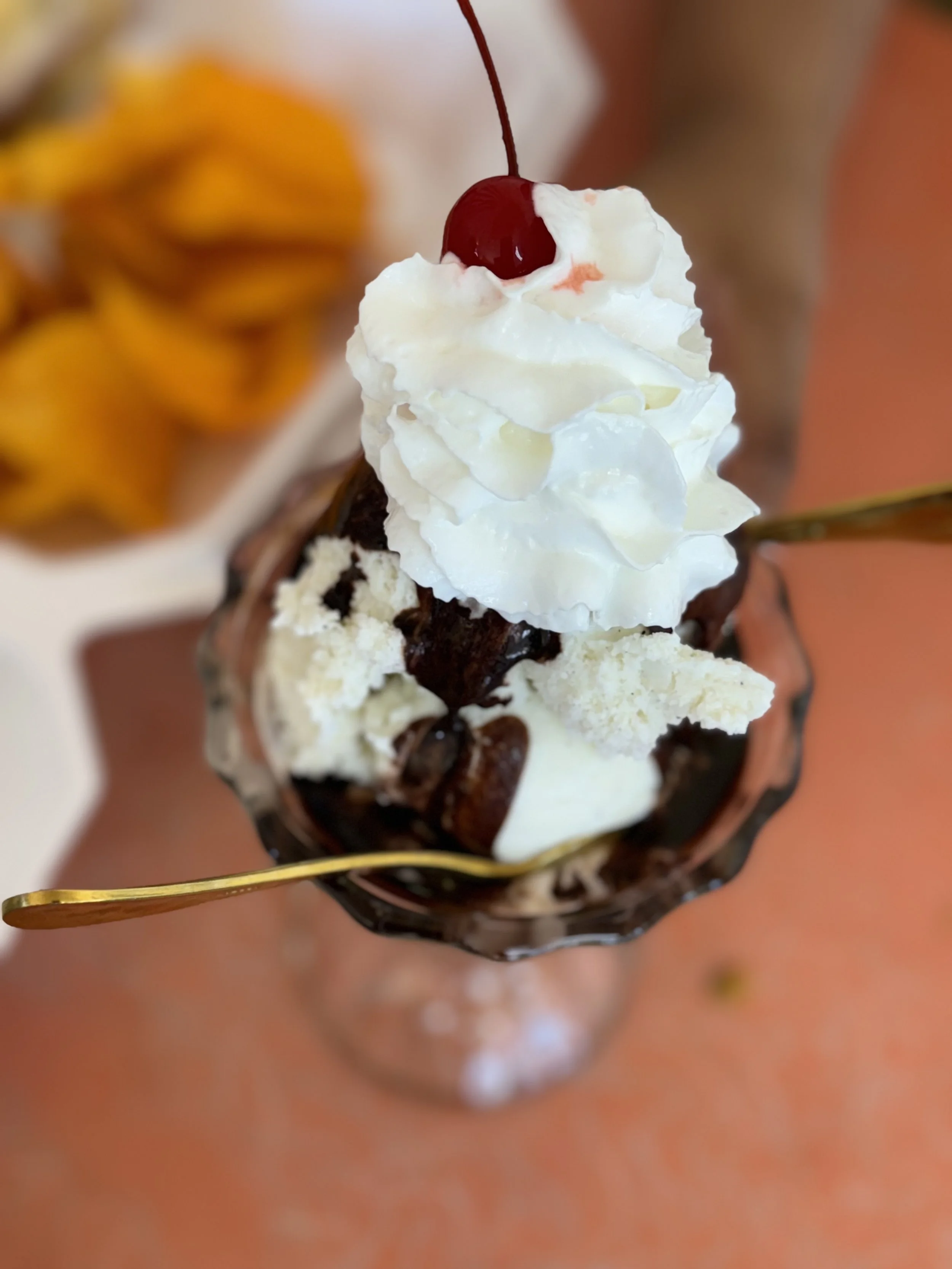 Chocolate sundae with whipped cream, cherry on top, in a glass, with a spoon, and blurred peach slices in the background.