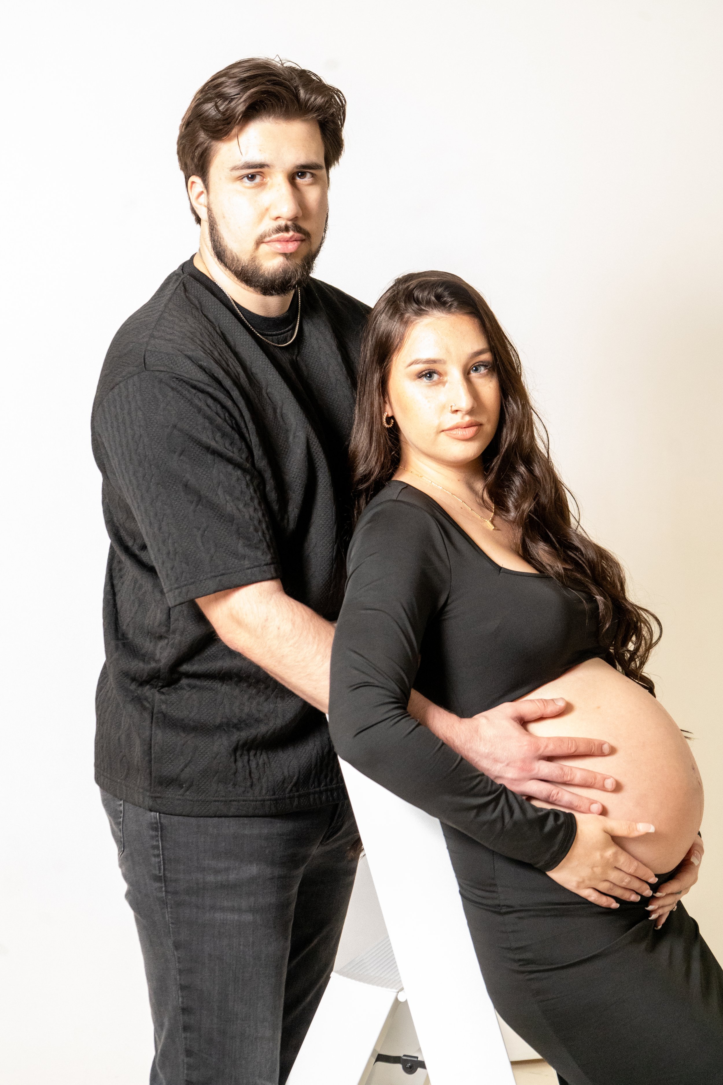 A pregnant woman with long, wavy brown hair and light skin, wearing a black long-sleeve dress, is being embraced from behind by a man with short, dark hair, a beard, and light skin, dressed in a black textured t-shirt and jeans, against a plain white