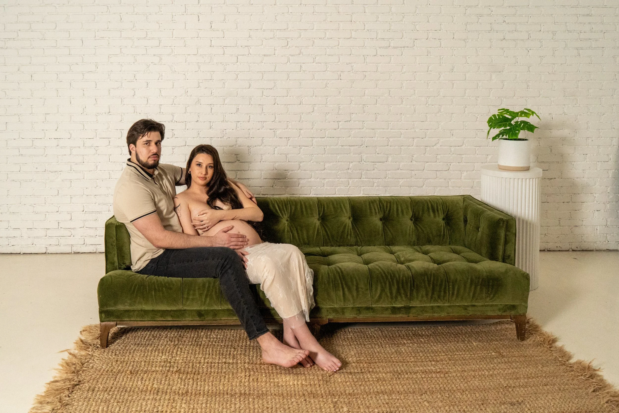 A man and a pregnant woman sitting on a green velvet couch in front of a white brick wall. The man has his arm around the woman, who is holding her pregnant belly. There is a potted plant on a white stand next to them.
