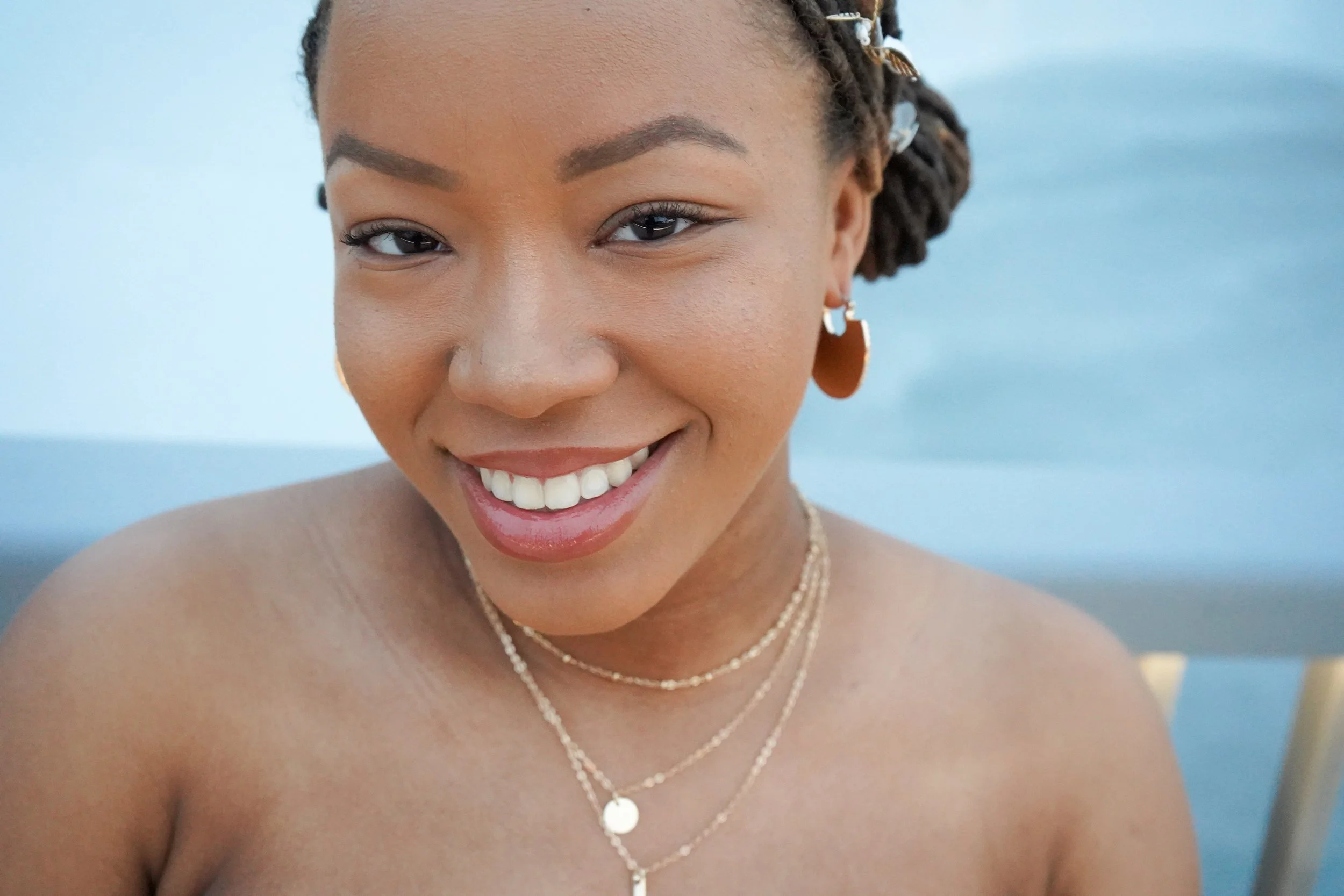 Close-up of a smiling woman with braided hair, earrings, and layered necklaces, outdoors with a blurred blue background.