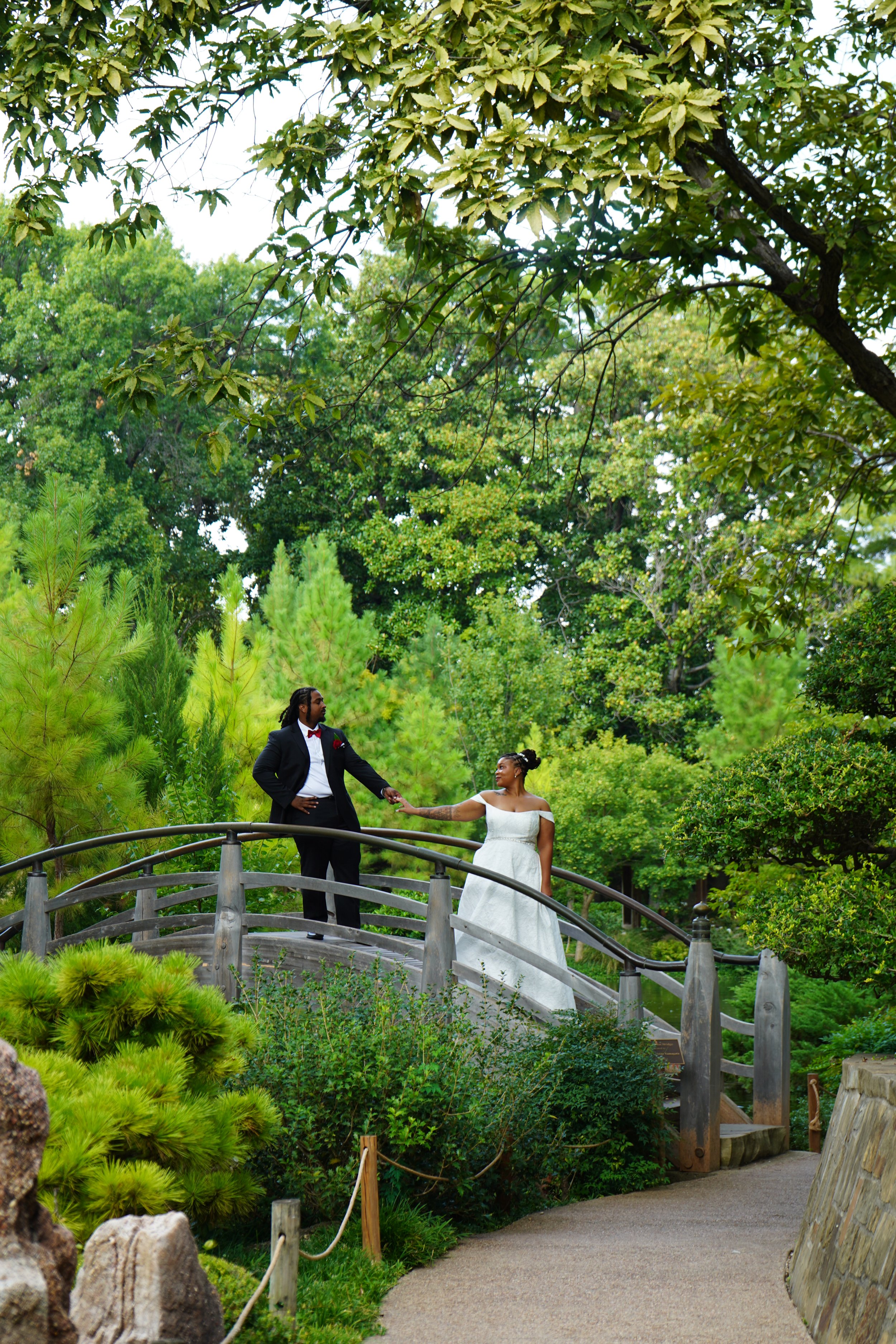 A couple dressed in formal wedding attire exploring a lush, green outdoor garden on a small wooden bridge. The groom is wearing a black tuxedo with a red bow tie, and the bride is in a white wedding dress.