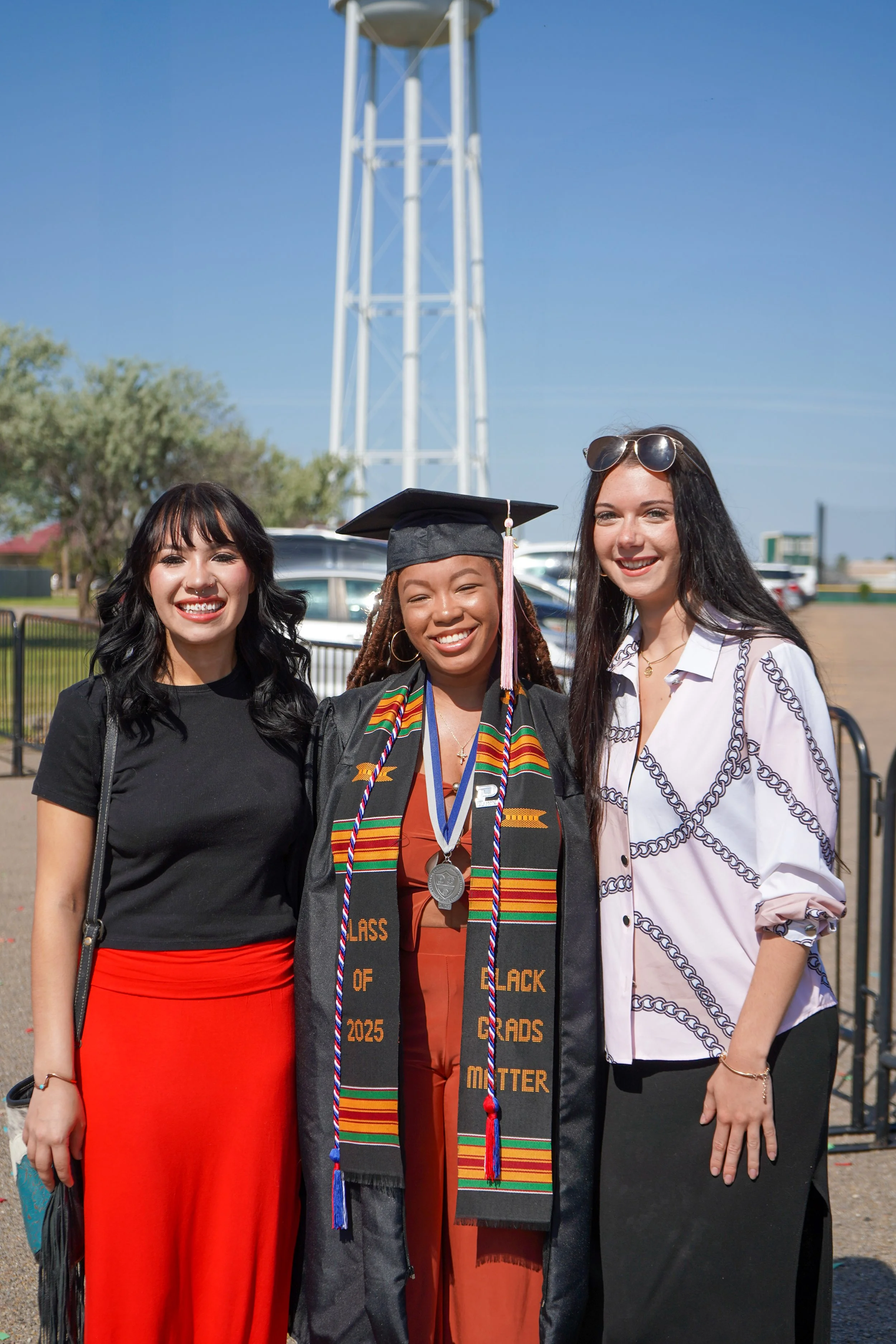 Three women celebrating graduation outdoors on a sunny day, with a tall water tower and cars in the background. The woman in the center is wearing a graduation cap and gown with a Kente cloth stole, medals, and cords. The women on either side are dressed in casual business attire.