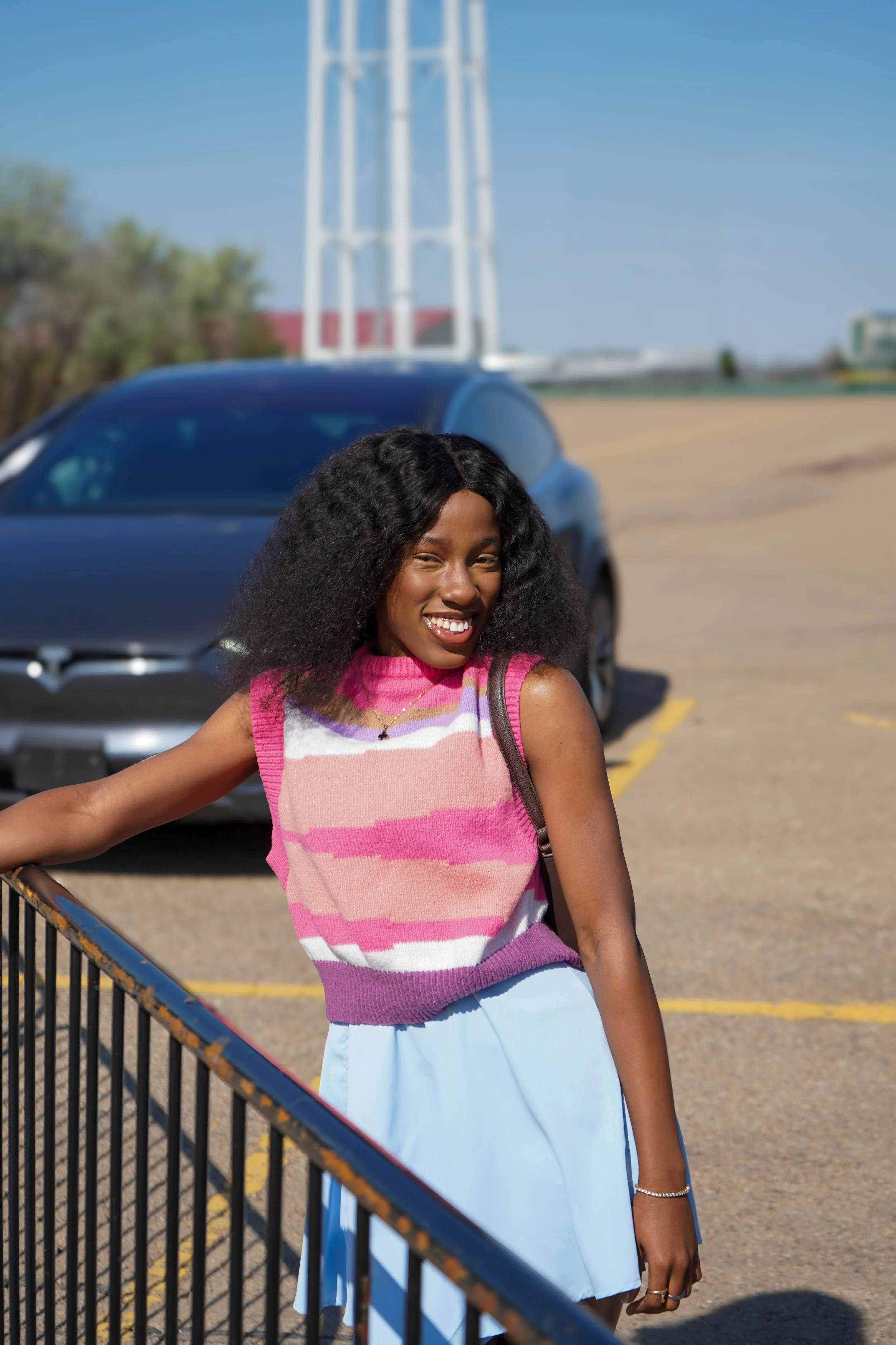 A woman smiling and leaning on a metal barrier in a parking lot with a black car and a windmill in the background on a clear, sunny day.