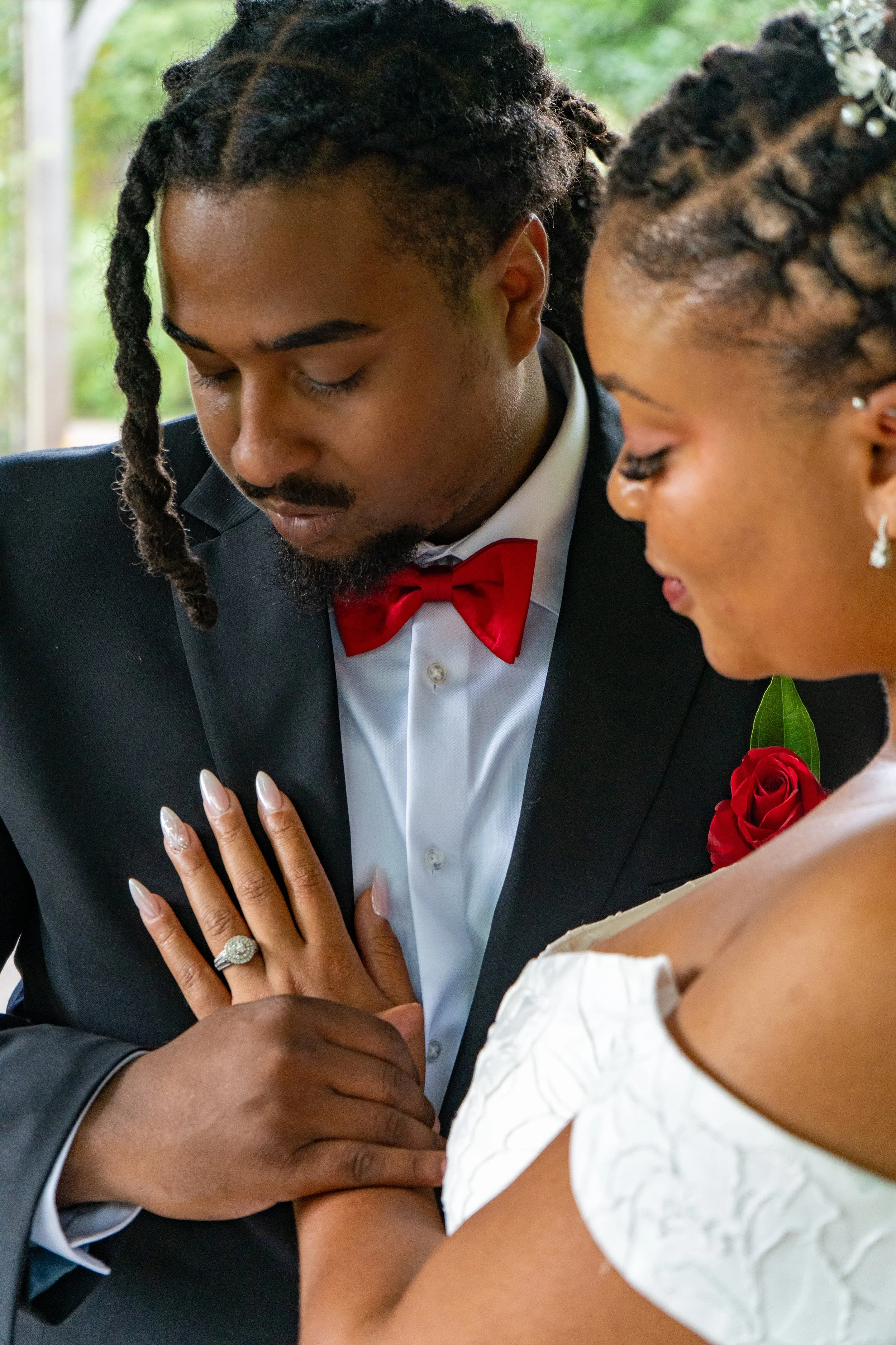 A close-up of a wedding couple, with the groom in a tuxedo with a red bow tie and the bride in an off-shoulder white dress, holding hands, displaying her wedding ring.